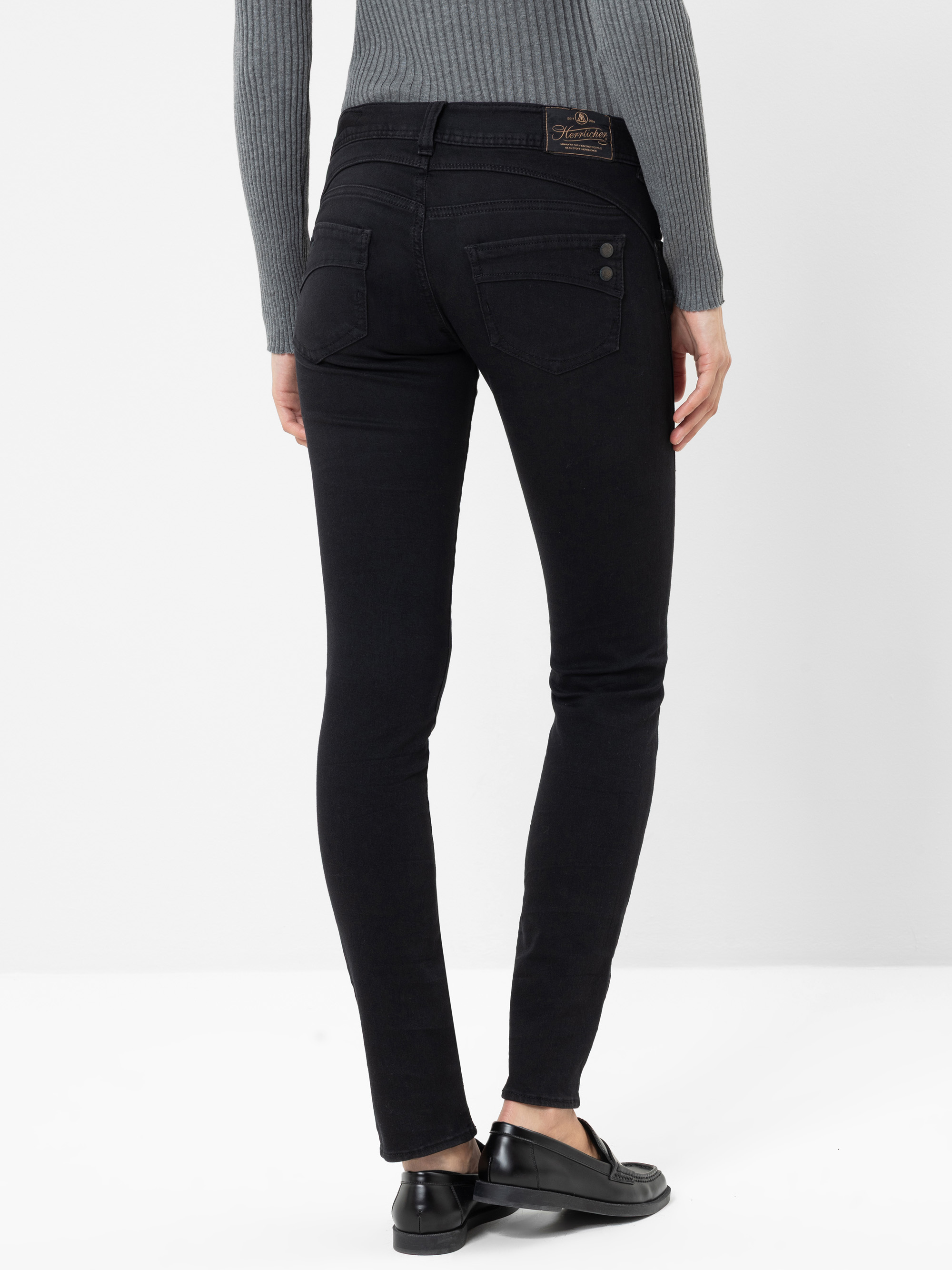 Eine Person, die einen grauen, gerippten Pullover, eine schwarze Skinny-Jeans mit Gesäßtaschen und schwarze Slipper trägt, steht mit dem Rücken zur Kamera vor einem schlichten weißen Hintergrund.