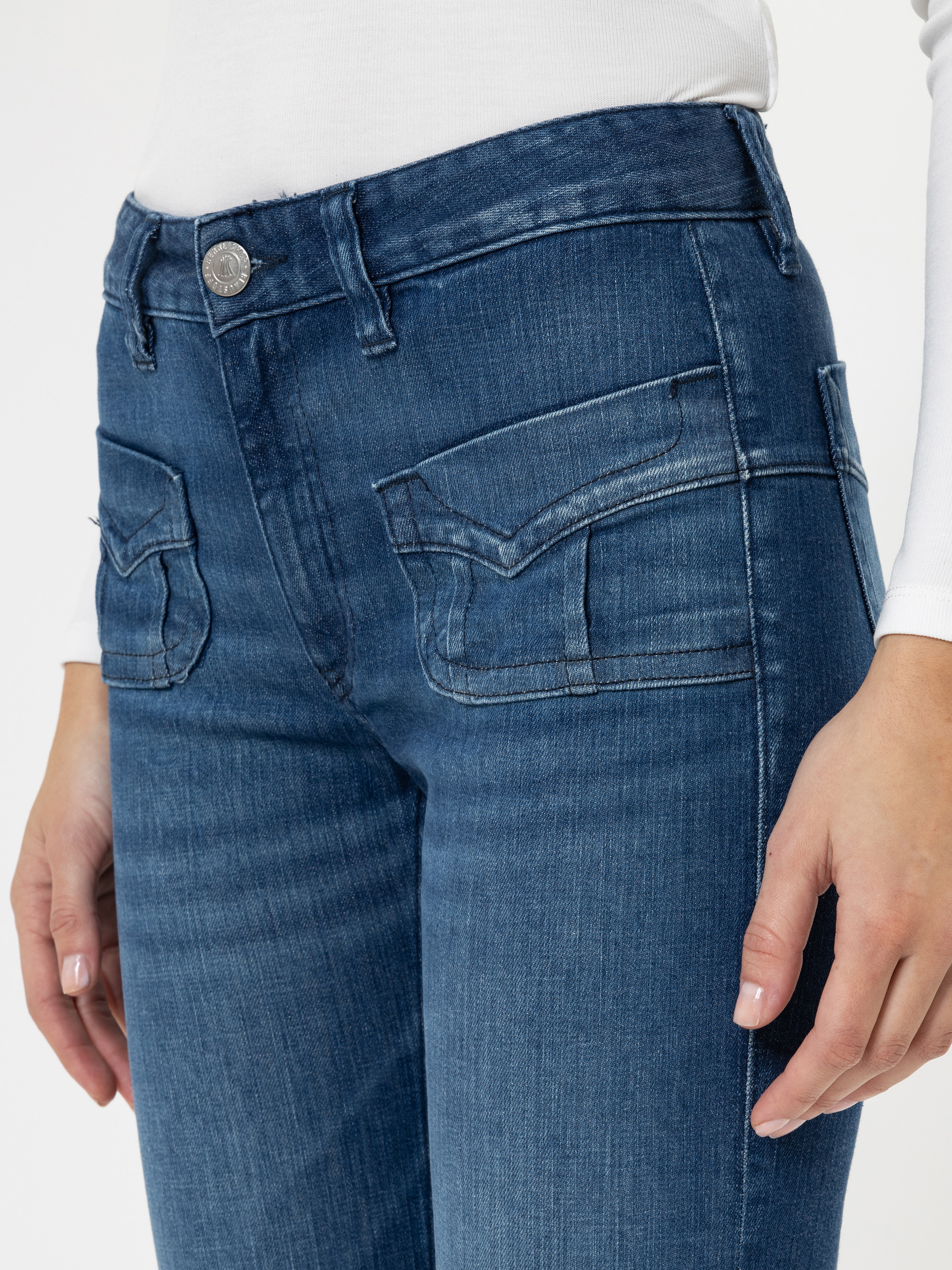 Eine Person, die eine blaue Jeans mit hoher Taille und Pattentaschen und ein weißes langärmeliges Oberteil trägt, wird von der Taille abwärts vor einem unifarbenen Hintergrund gezeigt.