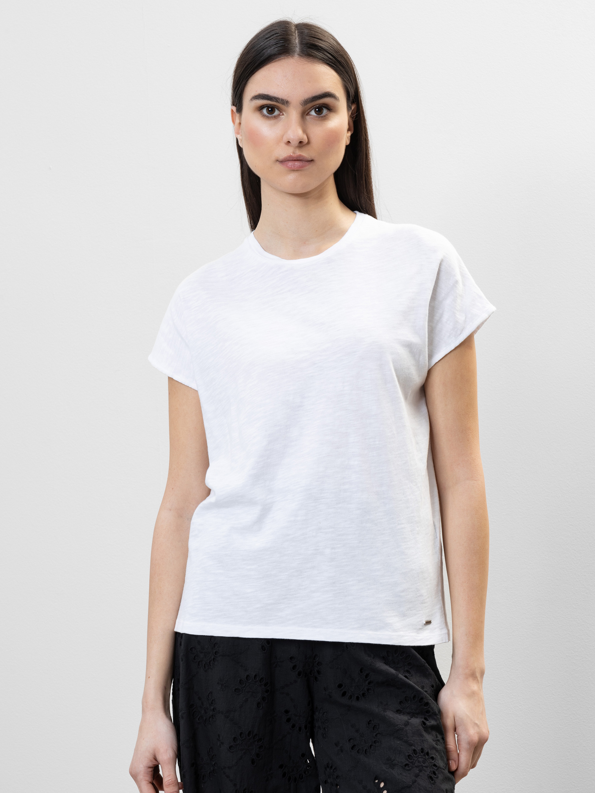 Eine Frau mit langem, glattem Haar trägt ein schlichtes weißes, kurzärmeliges T-Shirt und eine schwarz gemusterte Hose und steht vor einem schlichten, hellgrauen Hintergrund. Sie blickt direkt in die Kamera und hat einen neutralen Blick.