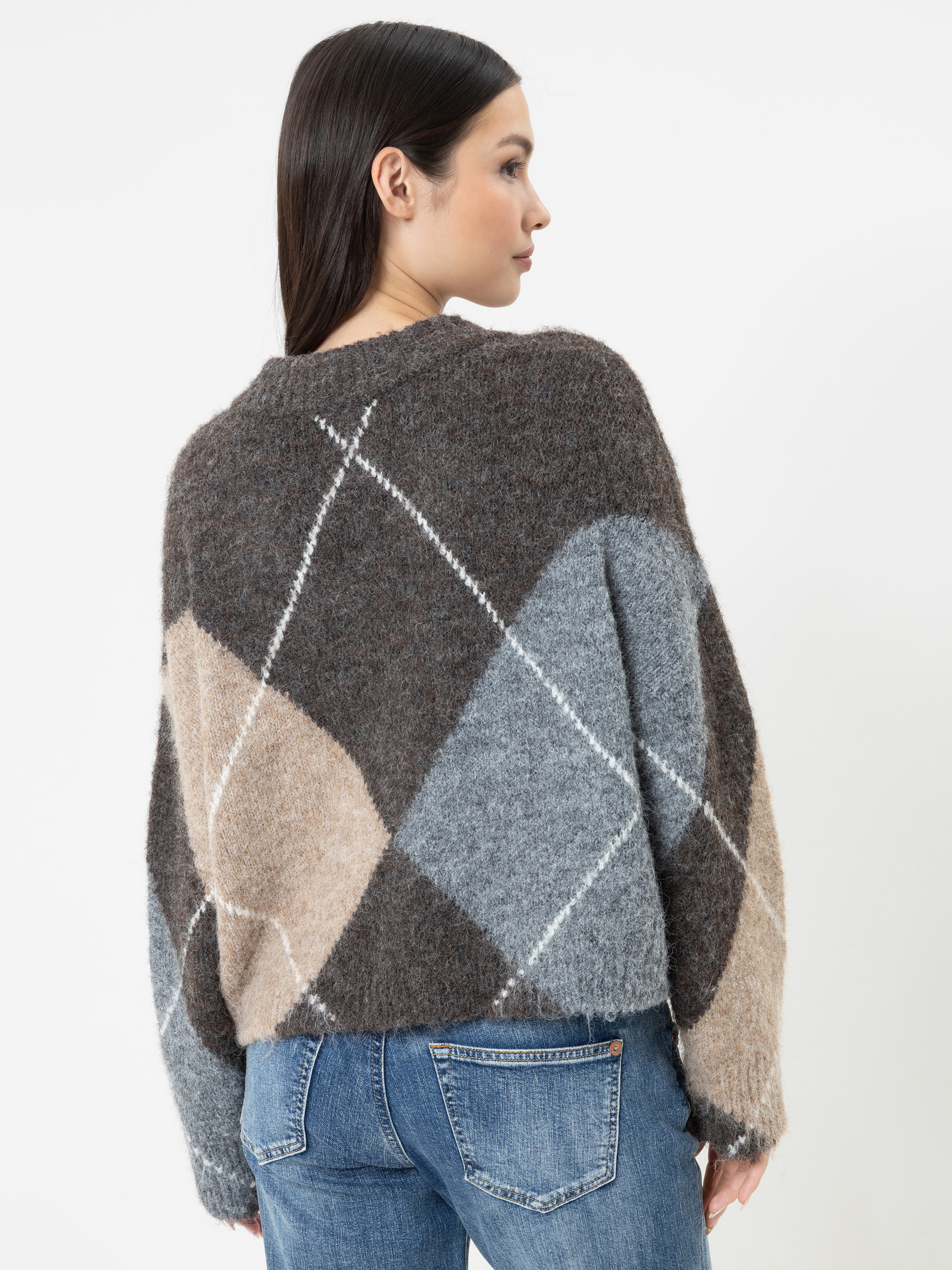 Eine Frau mit glattem, dunklem Haar trägt einen flauschigen, übergroßen Argyle-Pullover in Braun-, Grau- und Beigetönen, dazu eine blaue Jeans. Sie steht mit dem Rücken zur Kamera, sodass man die Rückseite des Pullovers vor einem schlichten weißen