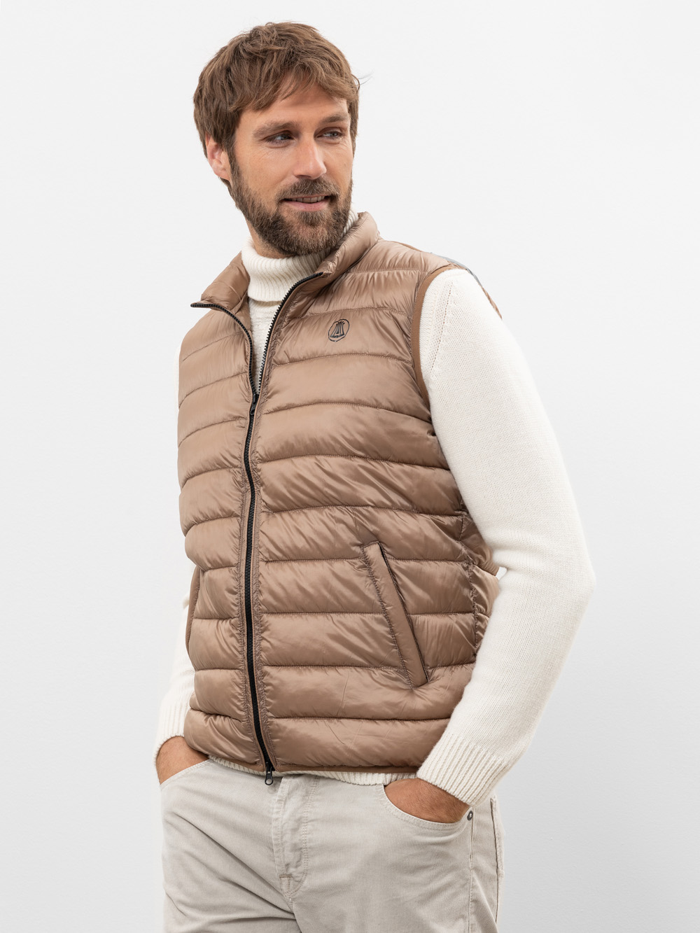 Ein Mann mit kurzen braunen Haaren und Bart trägt eine beigefarbene Kapuzenweste über einem weißen Rollkragenpullover und eine helle Hose. Er steht mit den Händen in den Taschen und blickt zur Seite vor einem schlichten weißen Hintergrund.