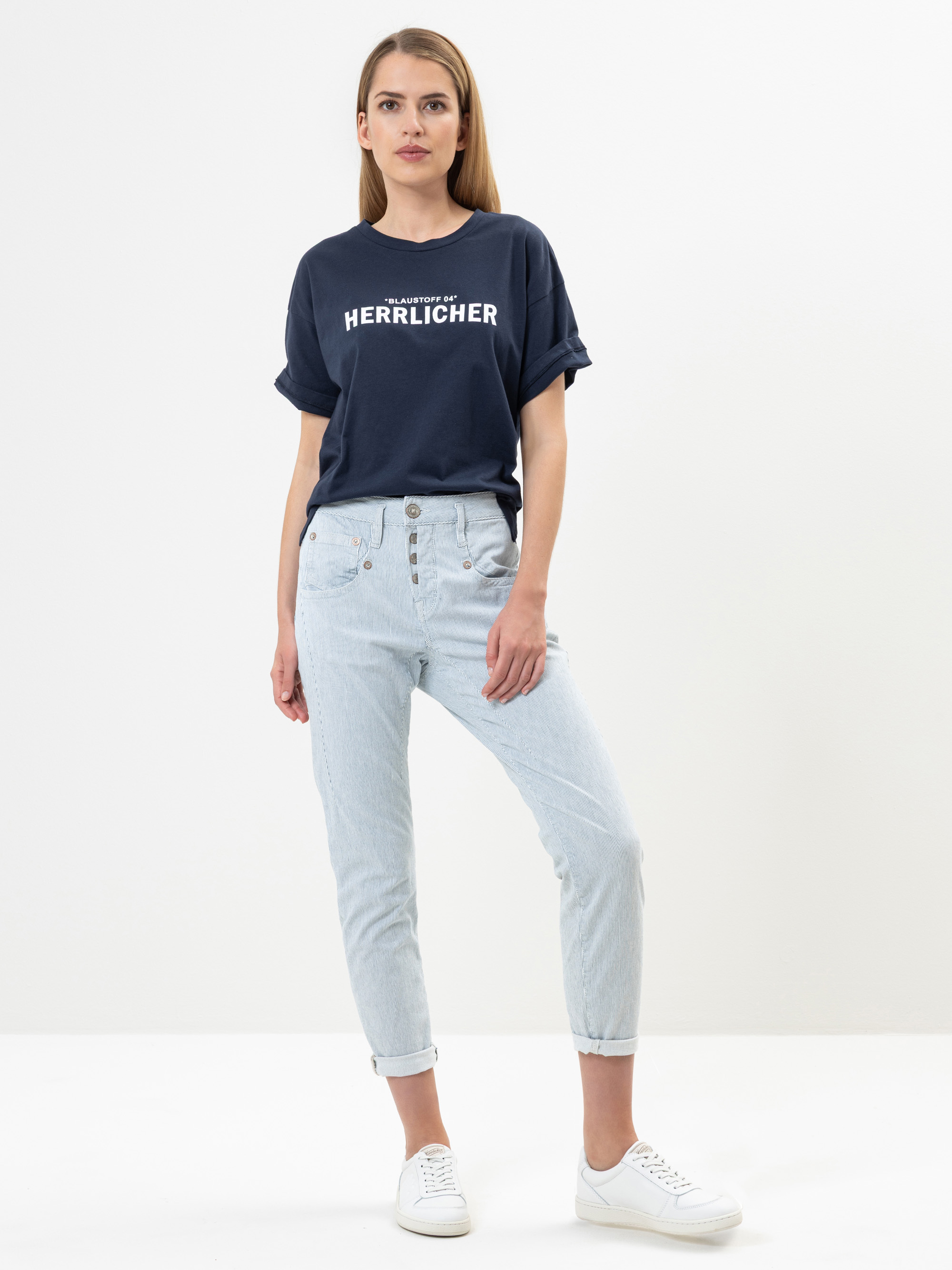 Eine Frau steht vor einem weißen Hintergrund und trägt ein marineblaues Herrlicher-T-Shirt, hellblaue Jeans mit Knöpfen und weiße Turnschuhe. Sie hat glattes, hellbraunes Haar und einen neutralen Gesichtsausdruck.