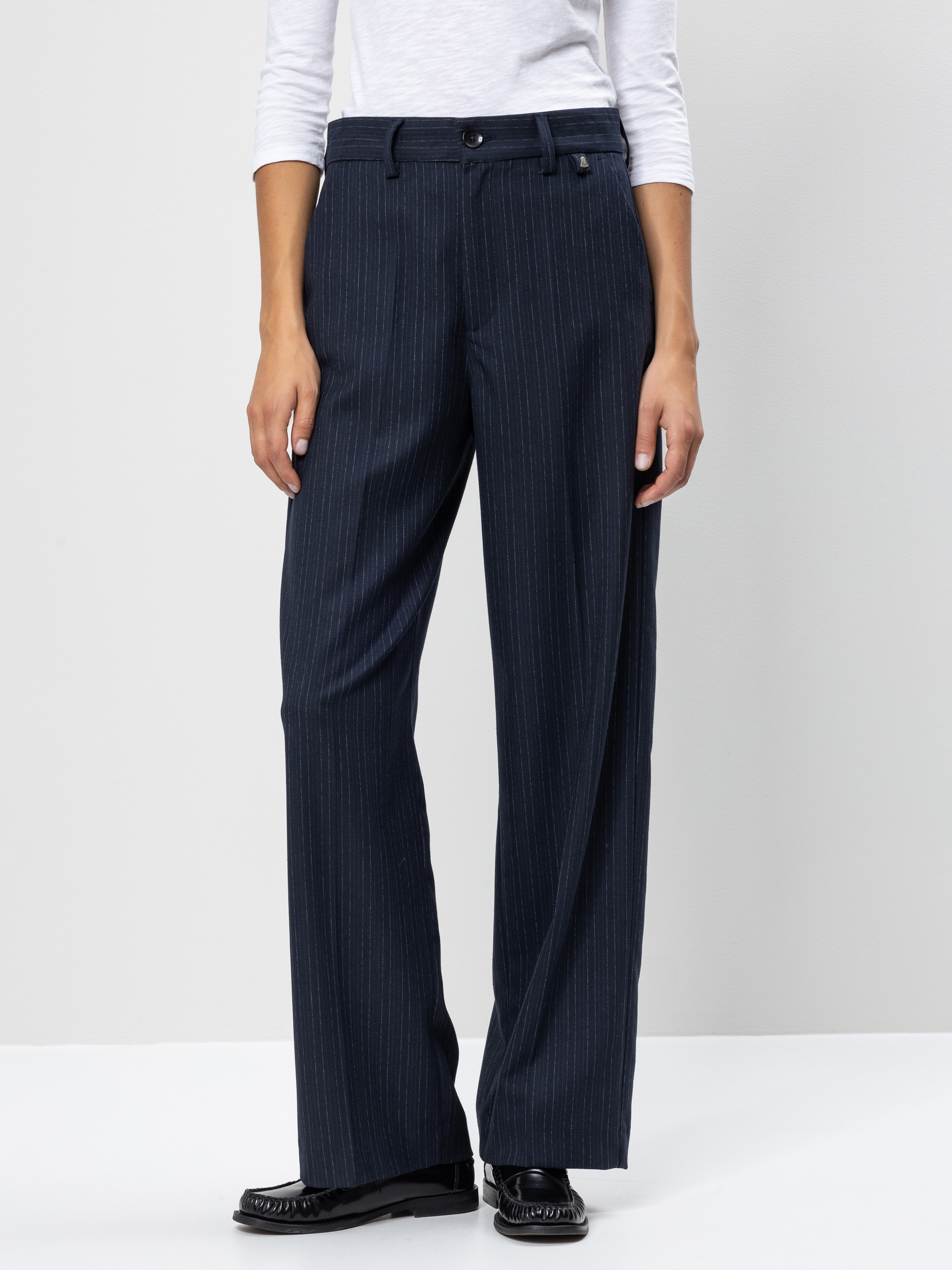 Eine Person, die eine marineblaue Nadelstreifenhose mit weitem Bein, ein weißes langärmeliges Oberteil und schwarze Halbschuhe trägt, steht vor einem schlichten hellgrauen Hintergrund.