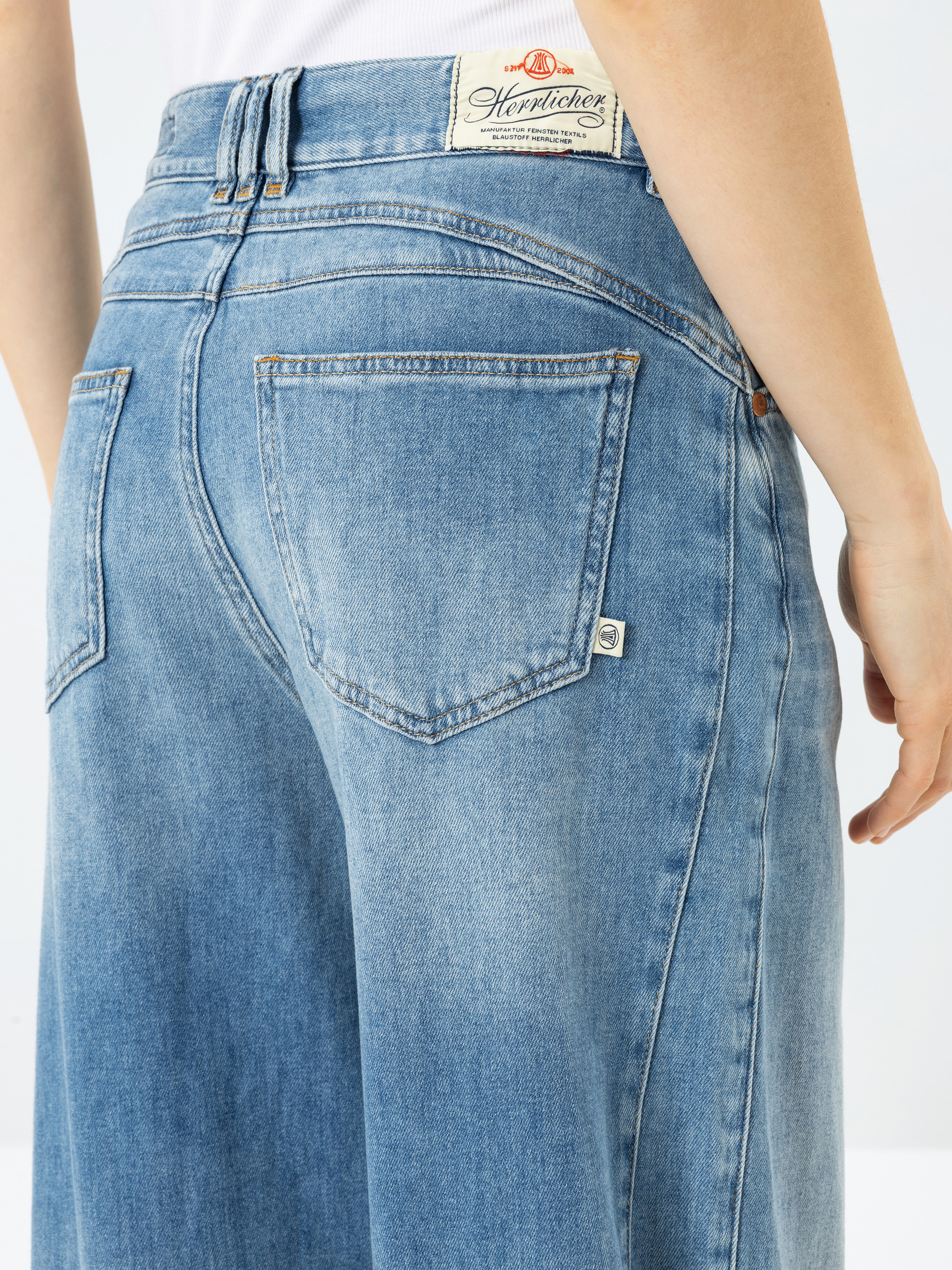 Nahaufnahme einer Person, die eine hellblaue Jeans mit weitem Bein trägt. Zu sehen sind die Gesäßtaschen, Nahtdetails und ein weißes Markenetikett "Hepitchick" am Bund. Der rechte Arm der Person ist an ihrer Seite zu sehen.