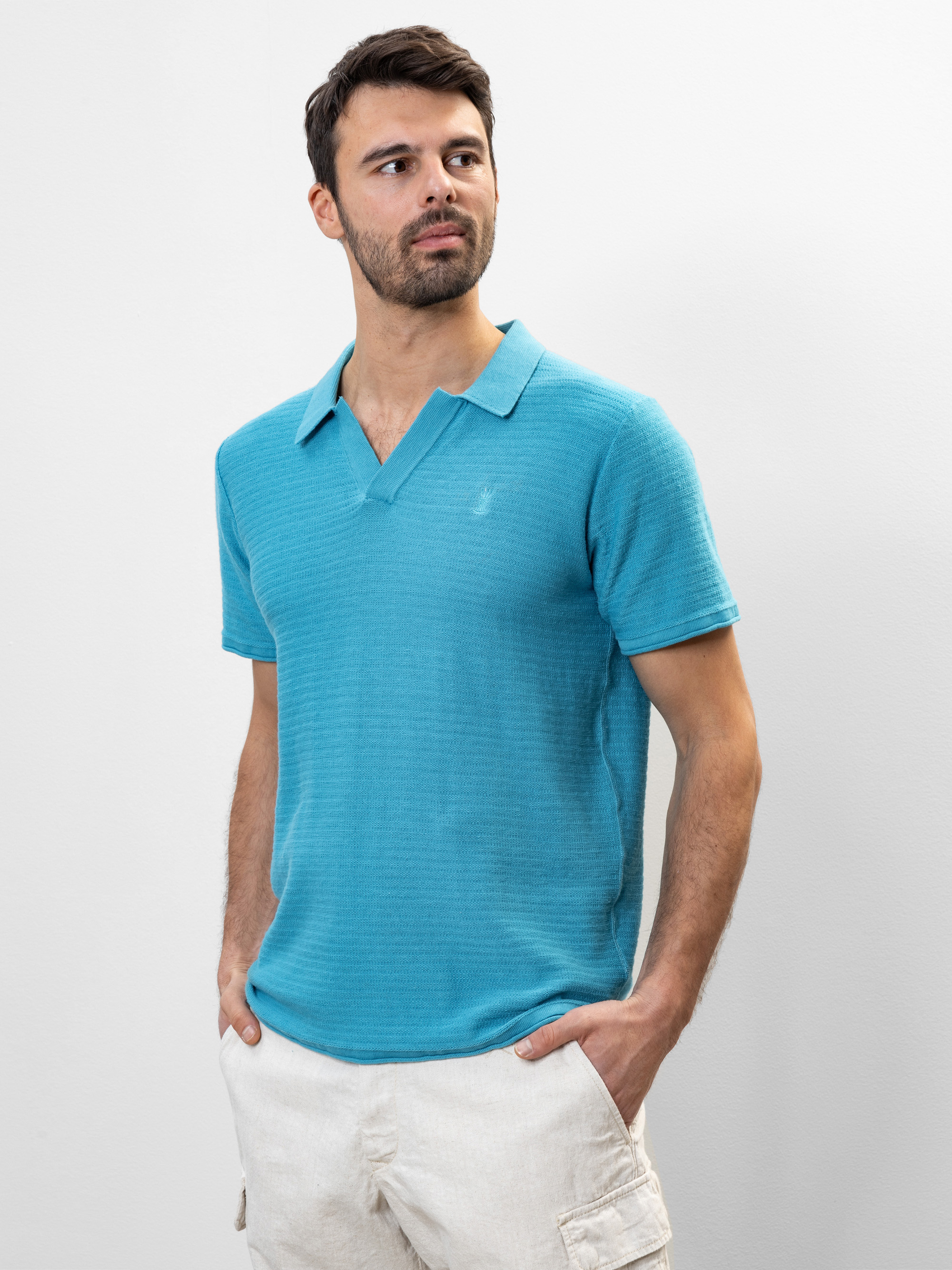 Ein Mann mit kurzen dunklen Haaren und einem Bart trägt ein hellblaues, kurzärmeliges Polohemd und beigefarbene Shorts. Er steht vor einem schlichten weißen Hintergrund, hat die Hände in den Taschen und schaut leicht zur Seite.