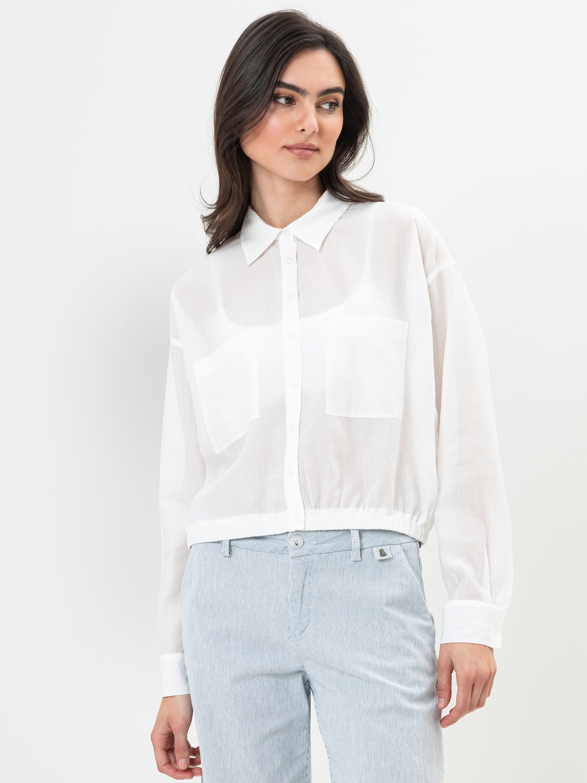 Eine Frau, die ein durchsichtiges weißes Hemd mit zwei Vordertaschen und eine hellblau gestreifte Hose trägt, steht vor einem schlichten weißen Hintergrund und schaut leicht nach links.