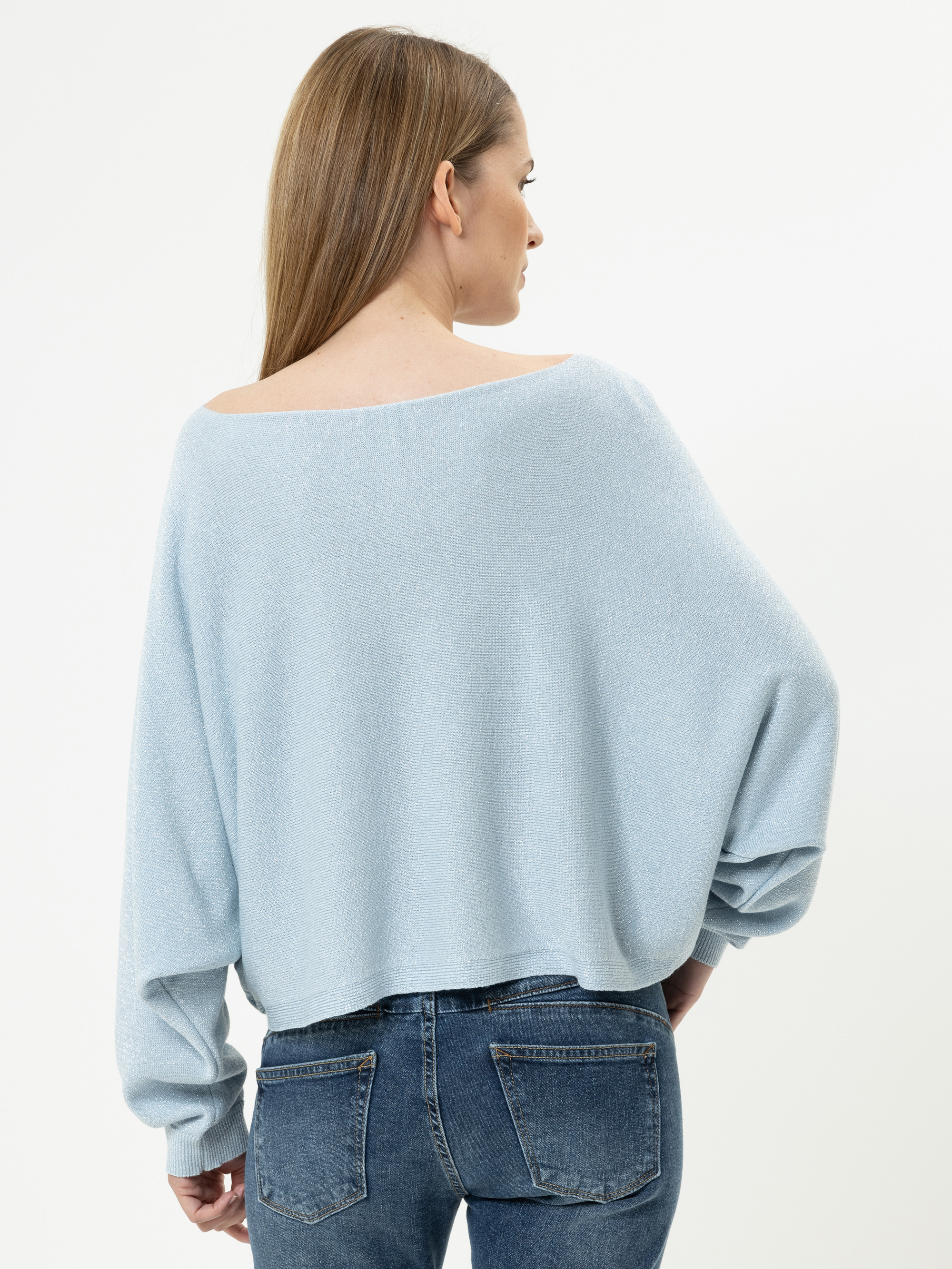 Eine Frau mit glattem, hellbraunem Haar trägt einen lockeren, hellblauen Pullover und blaue Jeans und steht mit dem Rücken zur Kamera vor einem schlichten weißen Hintergrund.