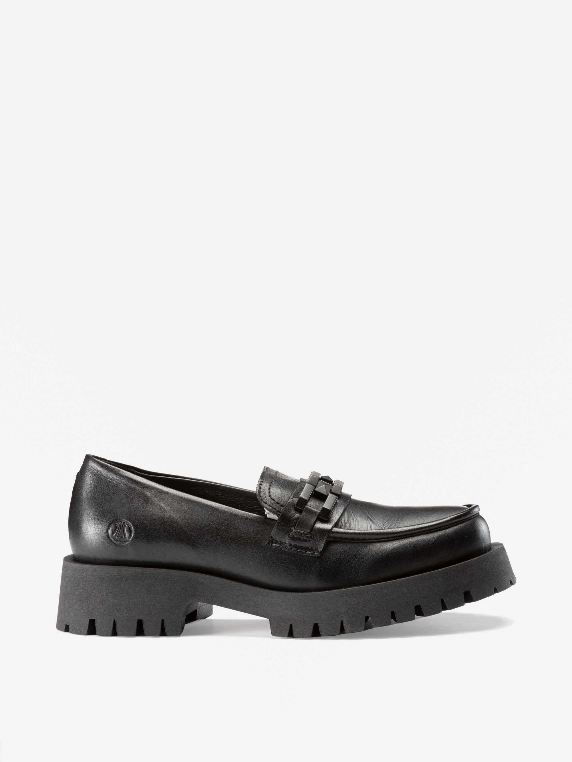 Ein schwarzer Lederslipper mit einer klobigen, geriffelten Sohle und einem dekorativen Riemen auf der Oberseite, von der Seite gesehen vor einem schlichten weißen Hintergrund.