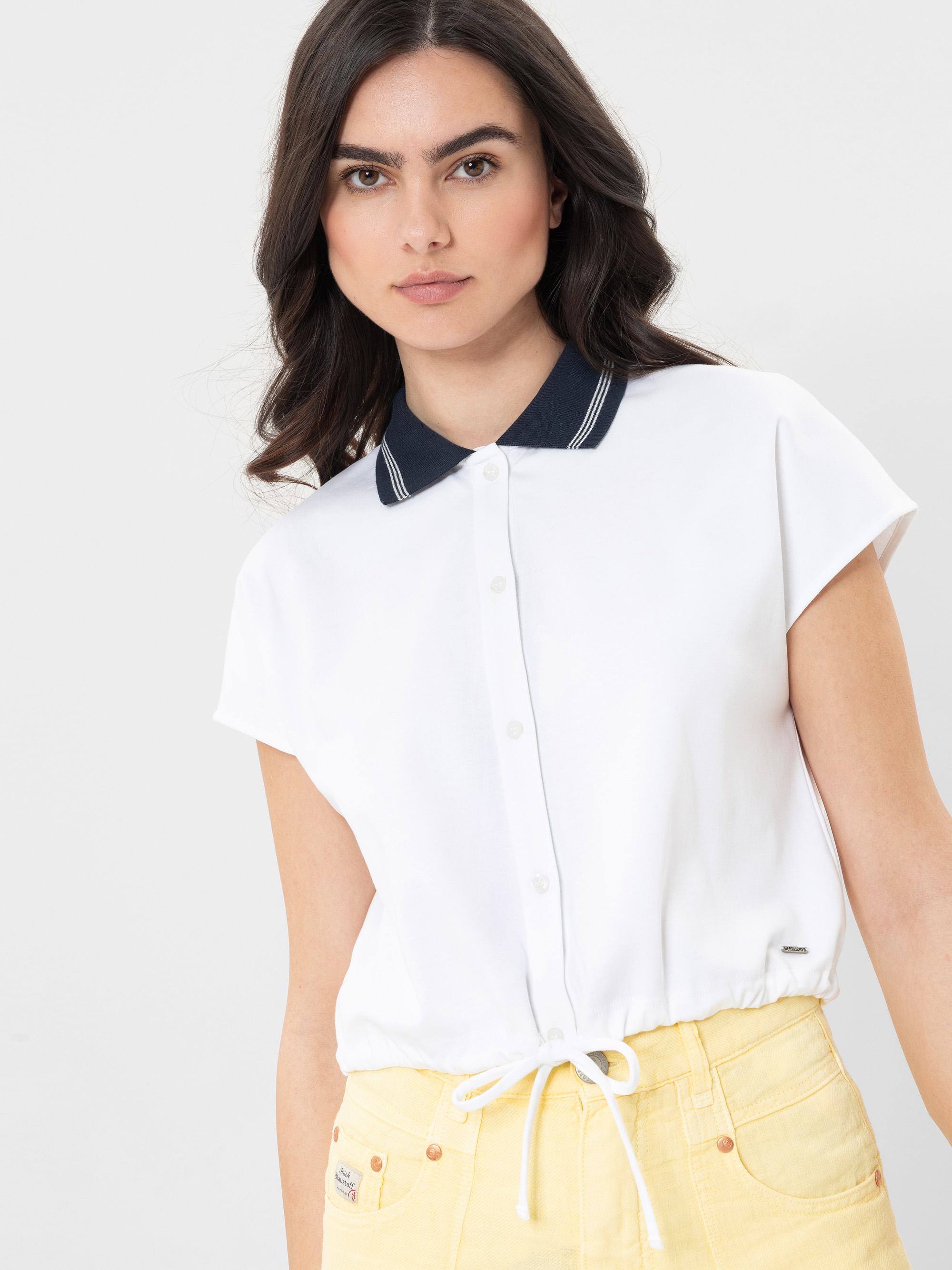 Eine Frau mit langen dunklen Haaren trägt ein kurzärmeliges weißes Button-up-Hemd mit marineblauem Kragen und eine gelbe Hose mit hoher Taille vor einem schlichten hellen Hintergrund.