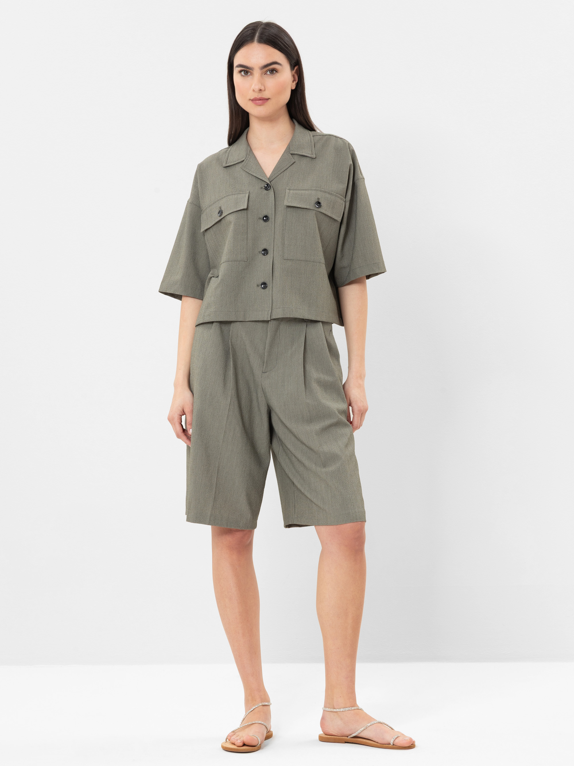 Eine Frau steht vor einem weißen Hintergrund und trägt ein locker sitzendes, kurzärmeliges, grau-grünes Button-up-Hemd mit Vordertaschen und dazu passende knielange Shorts. Sie hat langes glattes Haar und trägt flache Sandalen.
