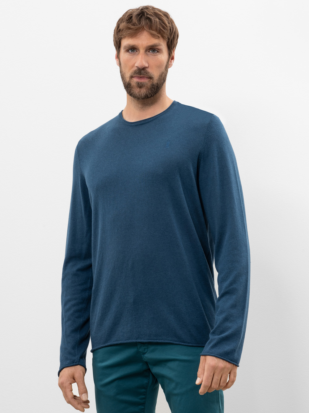 Ein Mann mit braunem Haar und Bart trägt ein langärmeliges, blaues T-Shirt mit Rundhalsausschnitt und eine blaugrüne Hose und steht vor einem schlichten weißen Hintergrund.