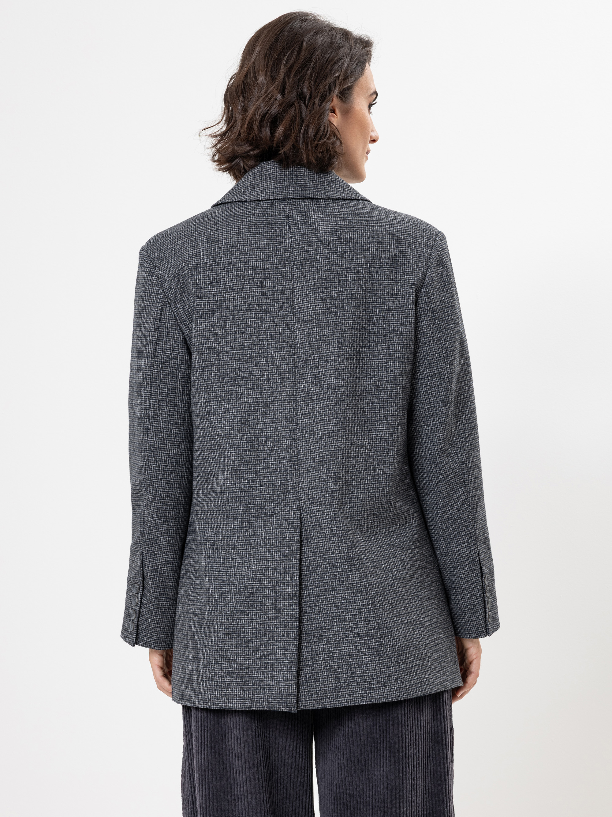 Eine Person mit kurzen braunen Haaren wird von hinten gezeigt, die einen grauen strukturierten Blazer und eine dunkle Hose trägt und vor einem schlichten weißen Hintergrund steht.