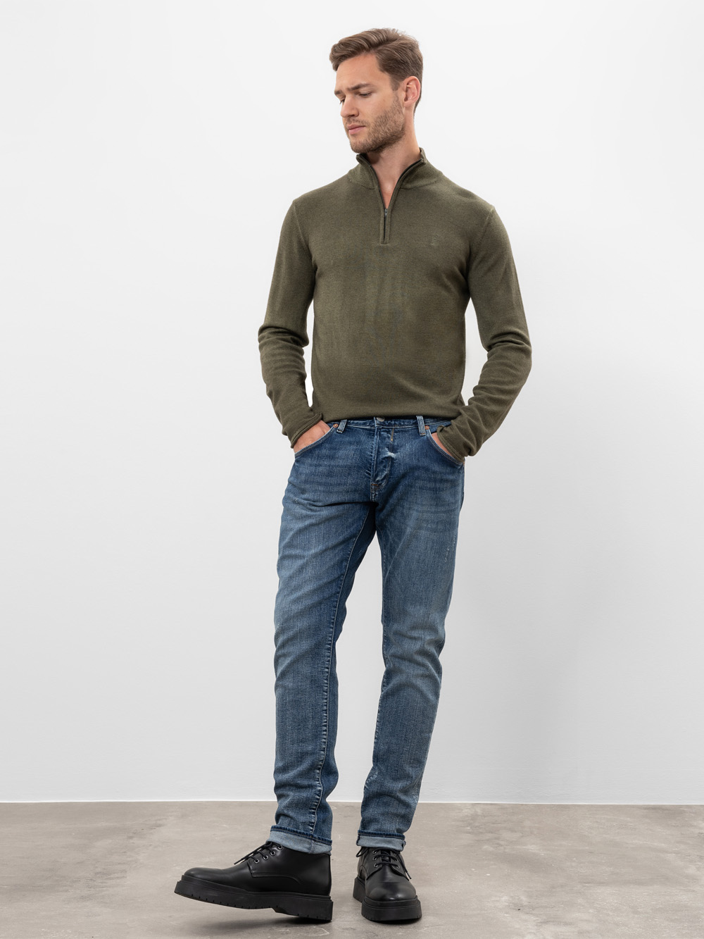 Ein Mann steht vor einem schlichten weißen Hintergrund, trägt einen grünen Pullover mit Viertelreißverschluss, blaue Jeans und schwarze Schuhe, hat die Hände in den Taschen und schaut leicht nach rechts.