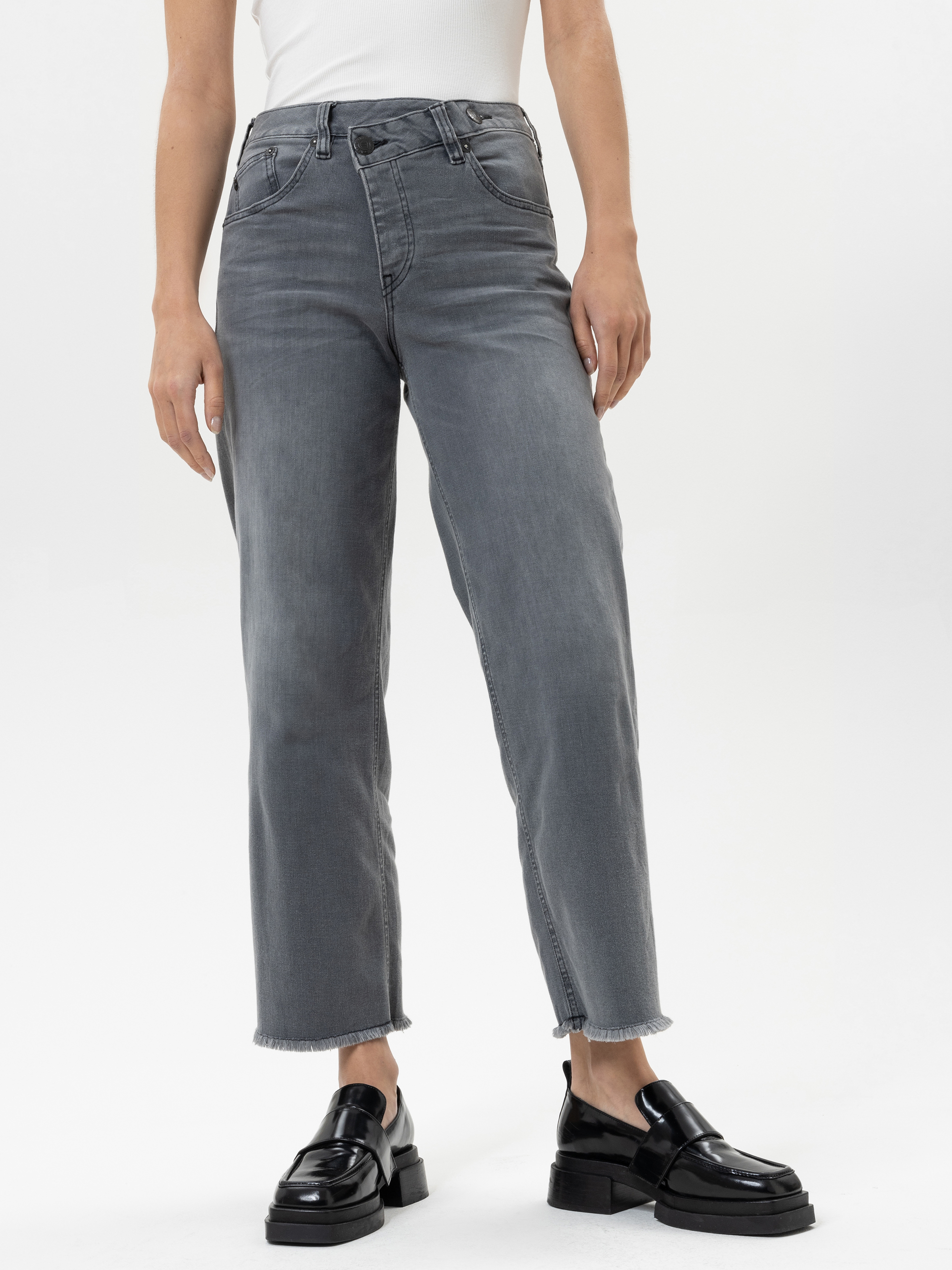 Eine Person, die eine graue Jeans mit hoher Taille und geradem Bein und rohem Saum, ein weißes Oberteil und schwarze Plateau-Slipper trägt, steht vor einem schlichten weißen Hintergrund.