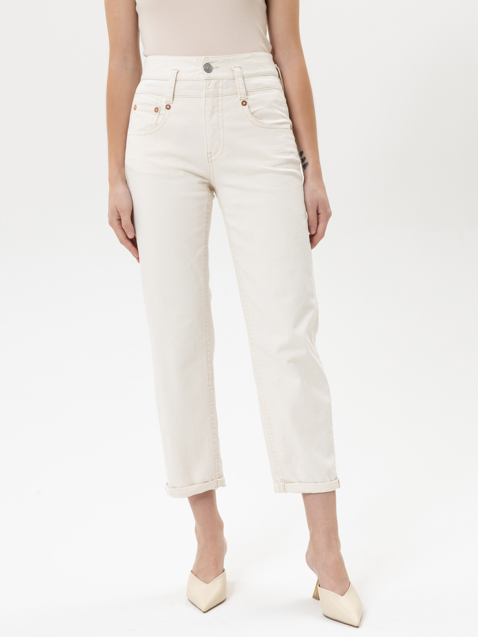 Eine Person steht vor einem weißen Hintergrund und trägt eine hoch taillierte, weiße Jeans mit geradem Bein und cremefarbene Schuhe mit spitzen Zehen. Ihre Arme liegen entspannt an der Seite und ihr Oberkörper ist teilweise außerhalb des Bildausschni