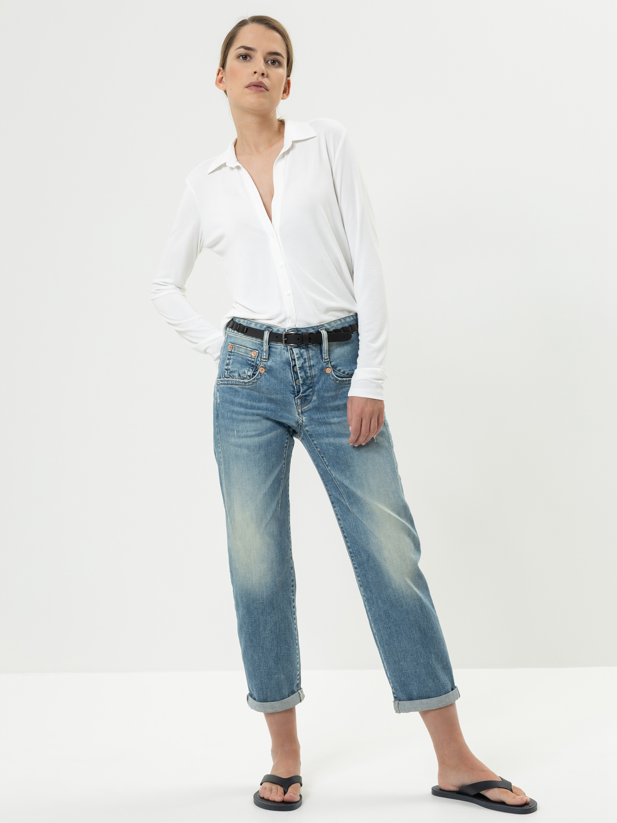 Eine Frau steht vor einem schlichten weißen Hintergrund, trägt ein weißes Button-up-Hemd, blaue Jeans mit Manschetten, einen schwarzen Gürtel und schwarze Flip-Flops, die Hände in den Taschen und einen neutralen Gesichtsausdruck.