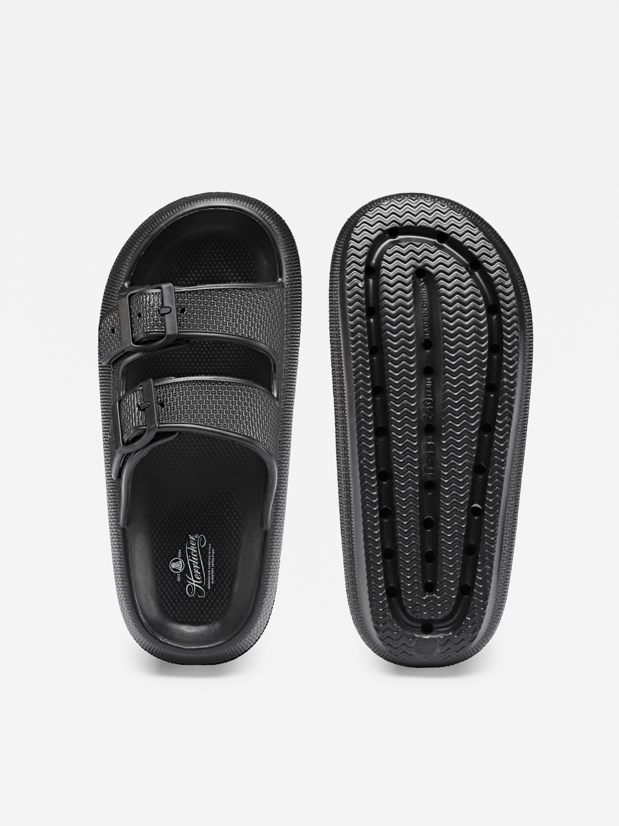 Ein Paar schwarze Gleitsandalen mit zwei Schnallen; eine Sandale ist von oben gezeigt, die andere zeigt die strukturierte Sohle mit Rillen und einem Zickzackmuster, alles auf weißem Hintergrund.