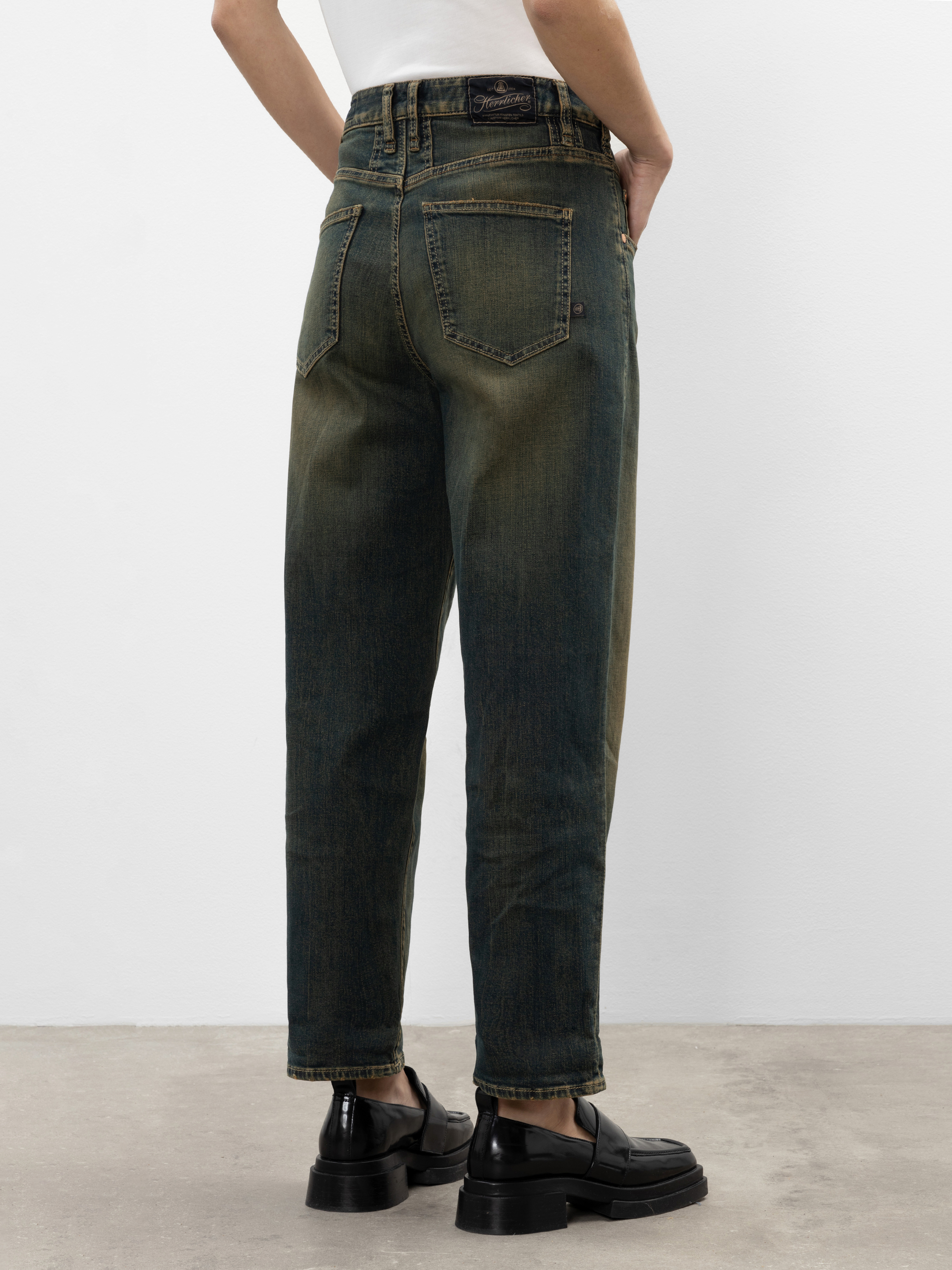 Eine Person in locker sitzenden, verblichenen Bluejeans und schwarzen Schuhen steht mit den Händen in den Hosentaschen, von der Kamera abgewandt, vor einem schlichten weißen Hintergrund.