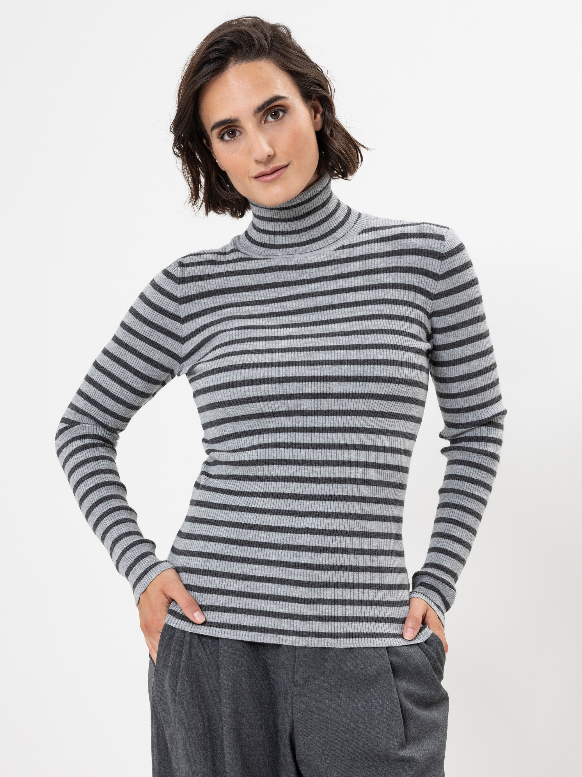 Eine Person mit kurzen braunen Haaren trägt einen grau-schwarz gestreiften Rollkragenpullover und eine graue Hose und steht mit den Händen in den Taschen vor einem schlichten weißen Hintergrund.