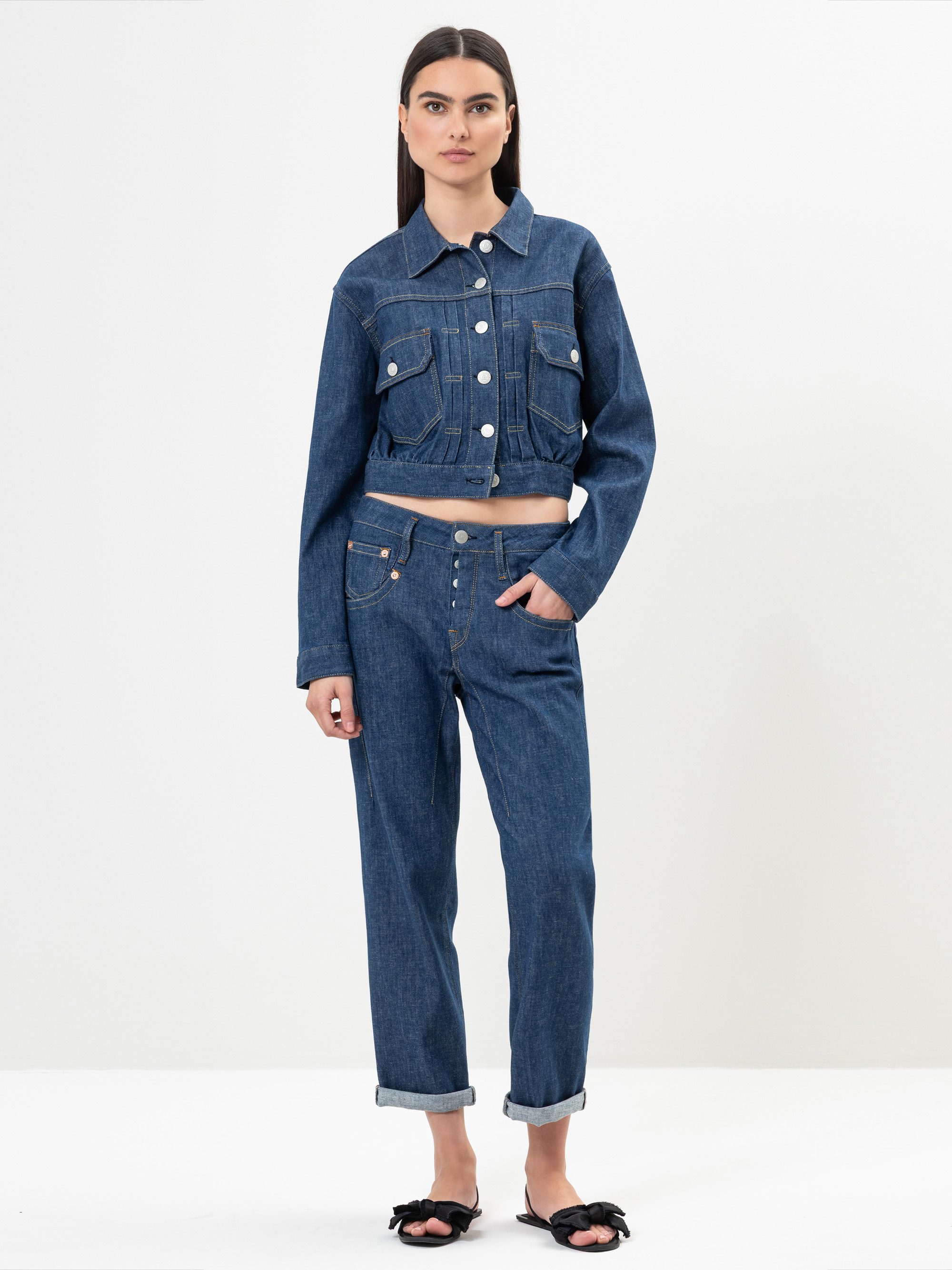 Eine Frau steht vor einem schlichten weißen Hintergrund und trägt eine Jeansjacke und passende Jeans mit hochgekrempelten Manschetten. Sie hat glattes dunkles Haar und trägt schwarze Sandalen mit Schleifen. Eine Hand steckt in ihrer Hosentasche; sie bl