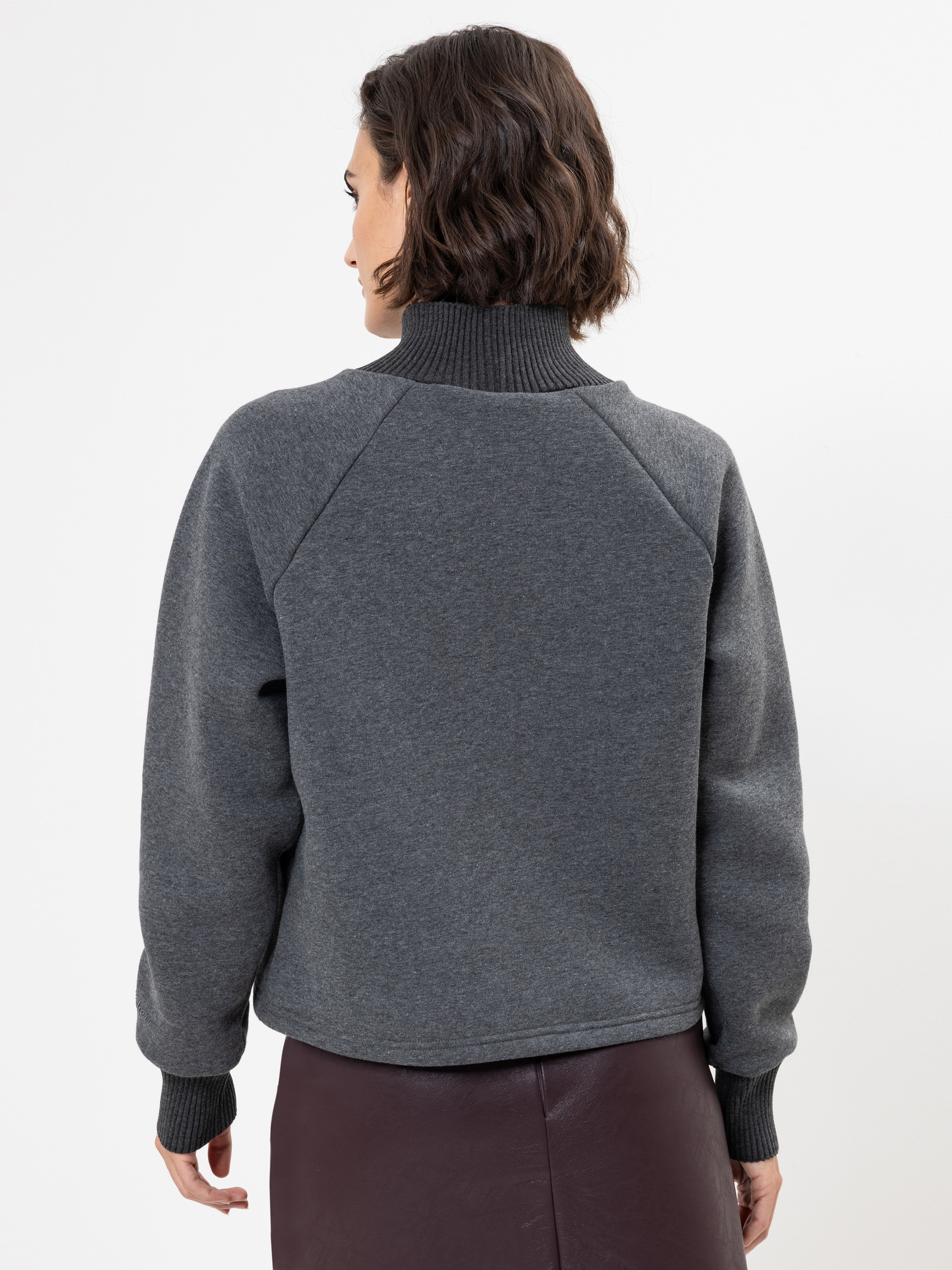 Eine Frau mit kurzen braunen Haaren steht mit dem Rücken zur Kamera, trägt einen dunkelgrauen, langärmeligen Pullover mit hohem Kragen und einen dunklen Rock vor einem schlichten weißen Hintergrund.