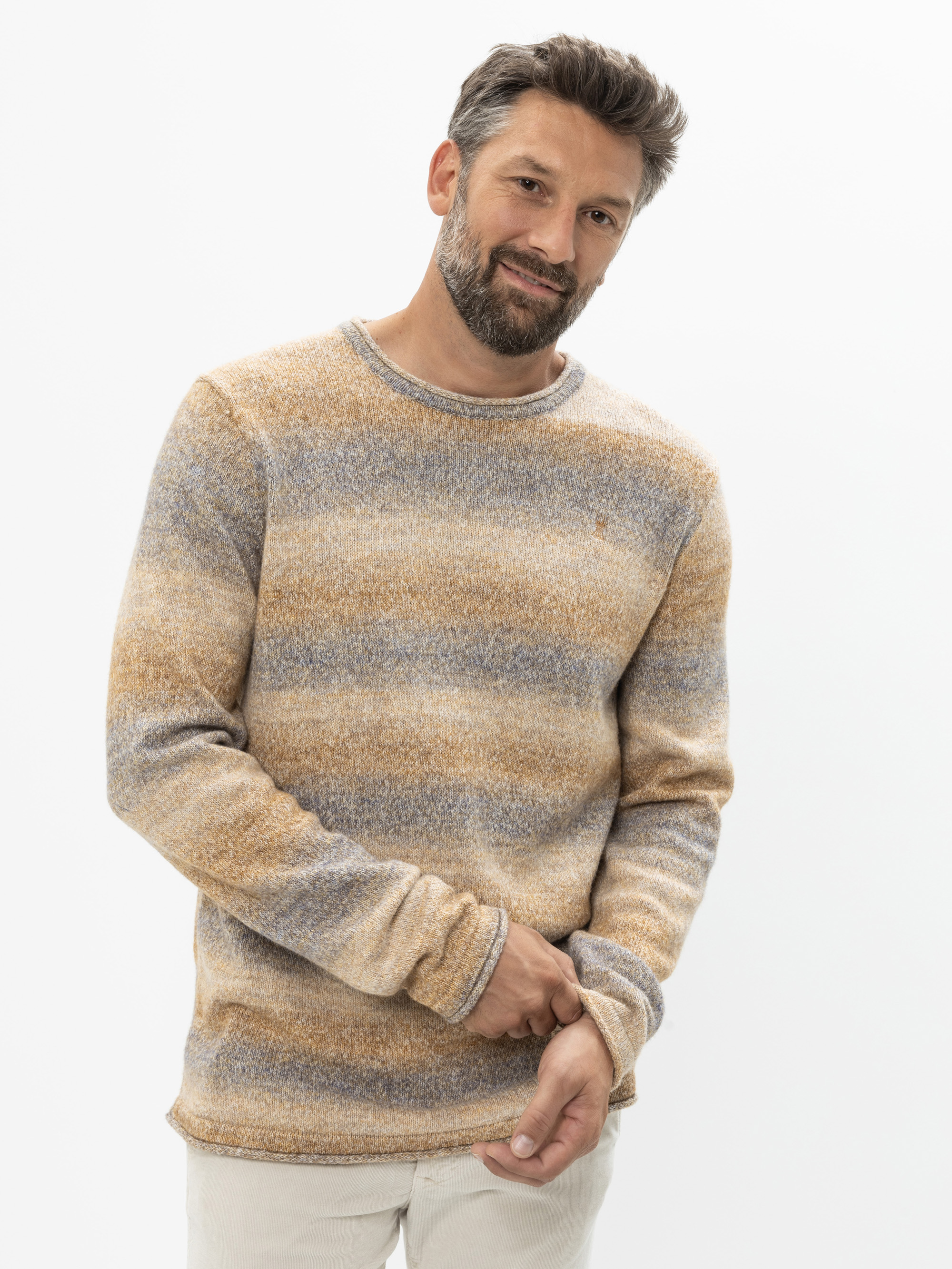 Ein Mann mit kurzen braunen Haaren und einem Bart, der einen beige-grau gestreiften Pullover und eine helle Hose trägt, steht vor einem weißen Hintergrund und lächelt leicht, während er mit einer Hand seinen Ärmel festhält.