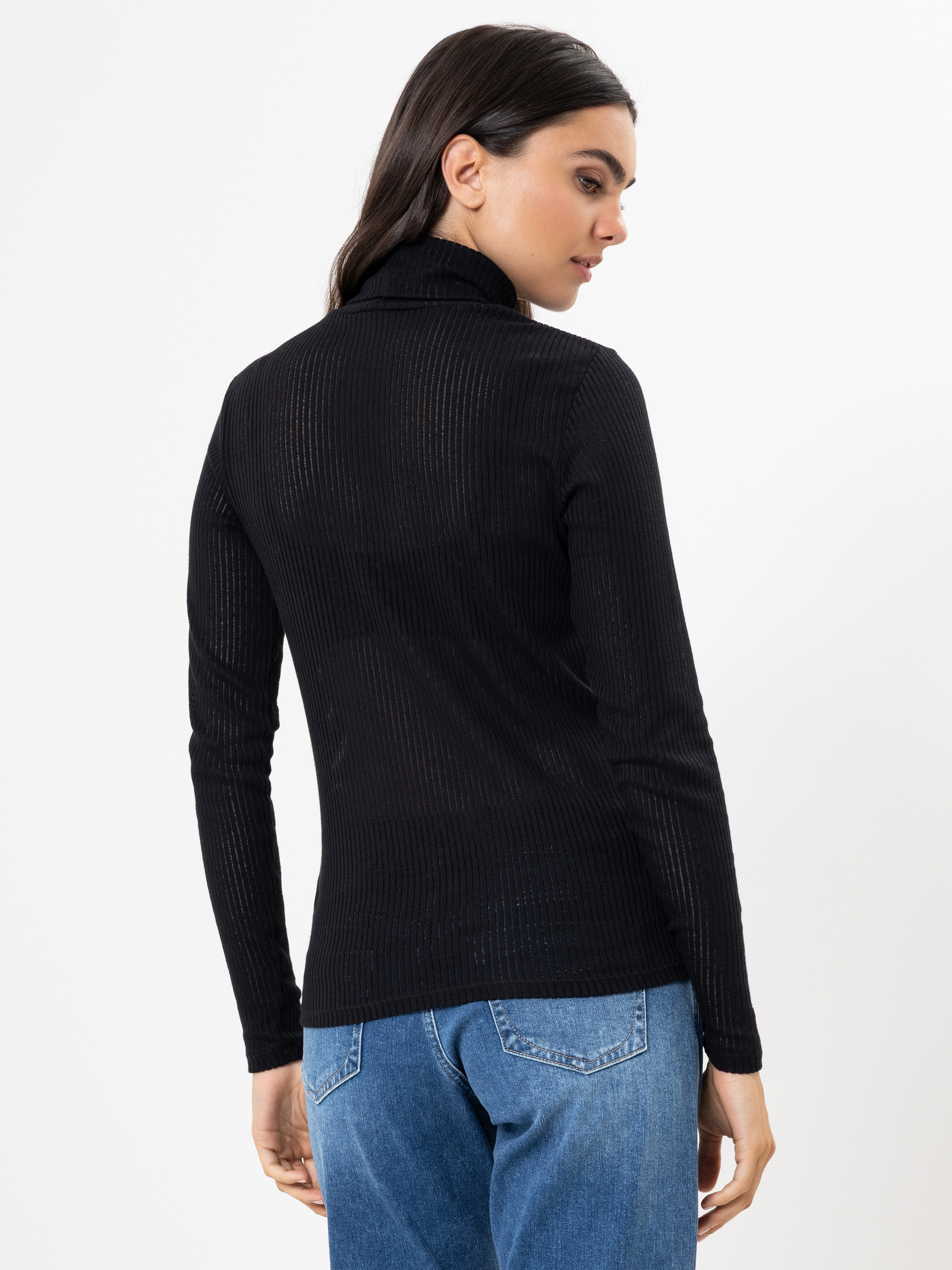 Eine Frau mit langen dunklen Haaren trägt ein schwarzes, langärmeliges, geripptes Rollkragenoberteil und blaue Jeans. Sie steht vor einem weißen Hintergrund und schaut über ihre Schulter.