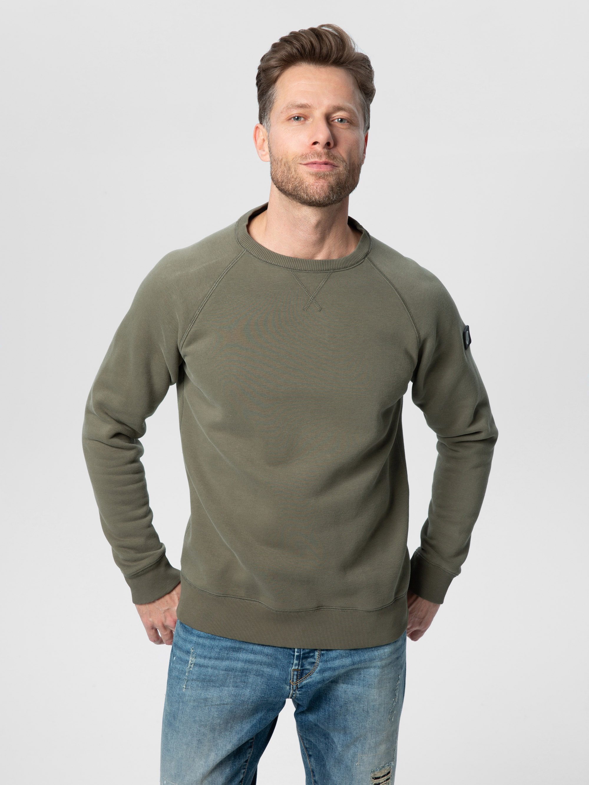 Ein Mann mit hellbraunem Haar und gestutztem Bart trägt ein olivgrünes Sweatshirt und blaue Jeans und steht mit den Händen in den Hüften vor einem schlichten hellgrauen Hintergrund.
