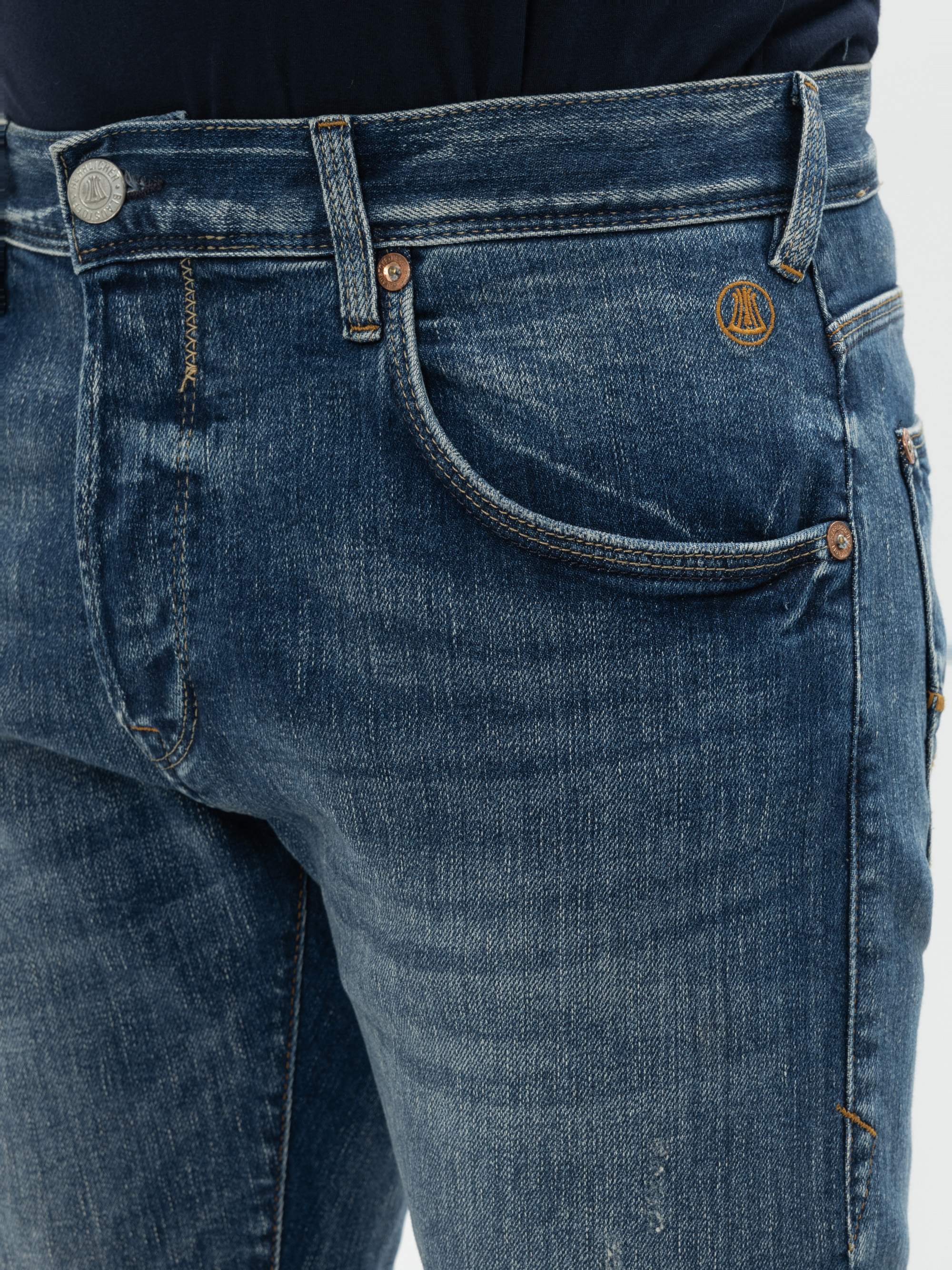 Nahaufnahme einer Person, die eine blaue Denim-Jeans mit einer sichtbaren Münztasche, Kupfernieten und einem kleinen gestickten runden Logo über der Tasche trägt. Die Jeans sieht leicht abgenutzt und verblasst aus.