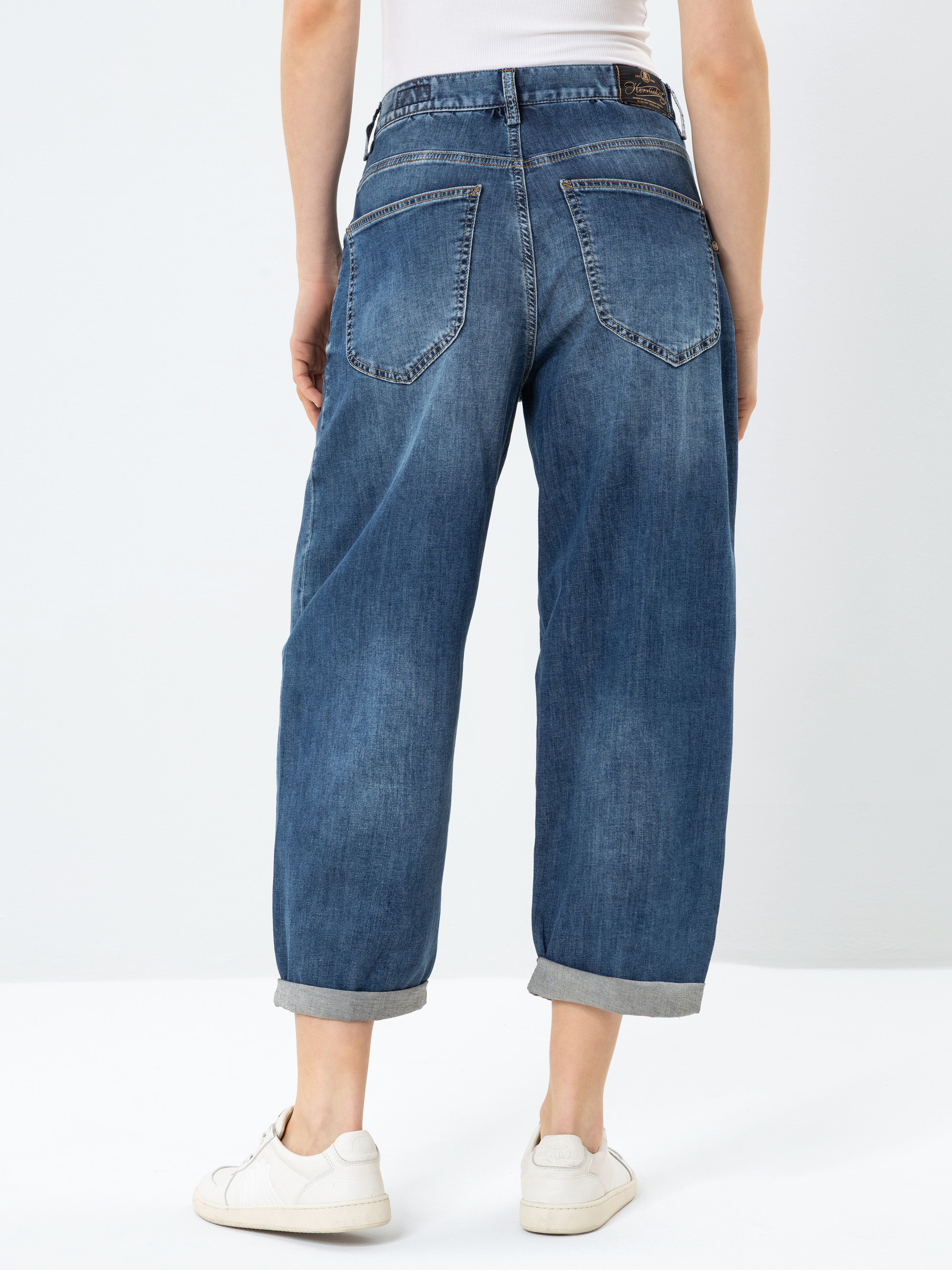 Person, die eine locker sitzende, mit Bündchen versehene blaue Jeans und weiße Turnschuhe trägt, von hinten vor einem schlichten hellen Hintergrund. Die Jeans hat zwei große Gesäßtaschen und wird mit einem weißen Oberteil kombiniert, das in die Hos