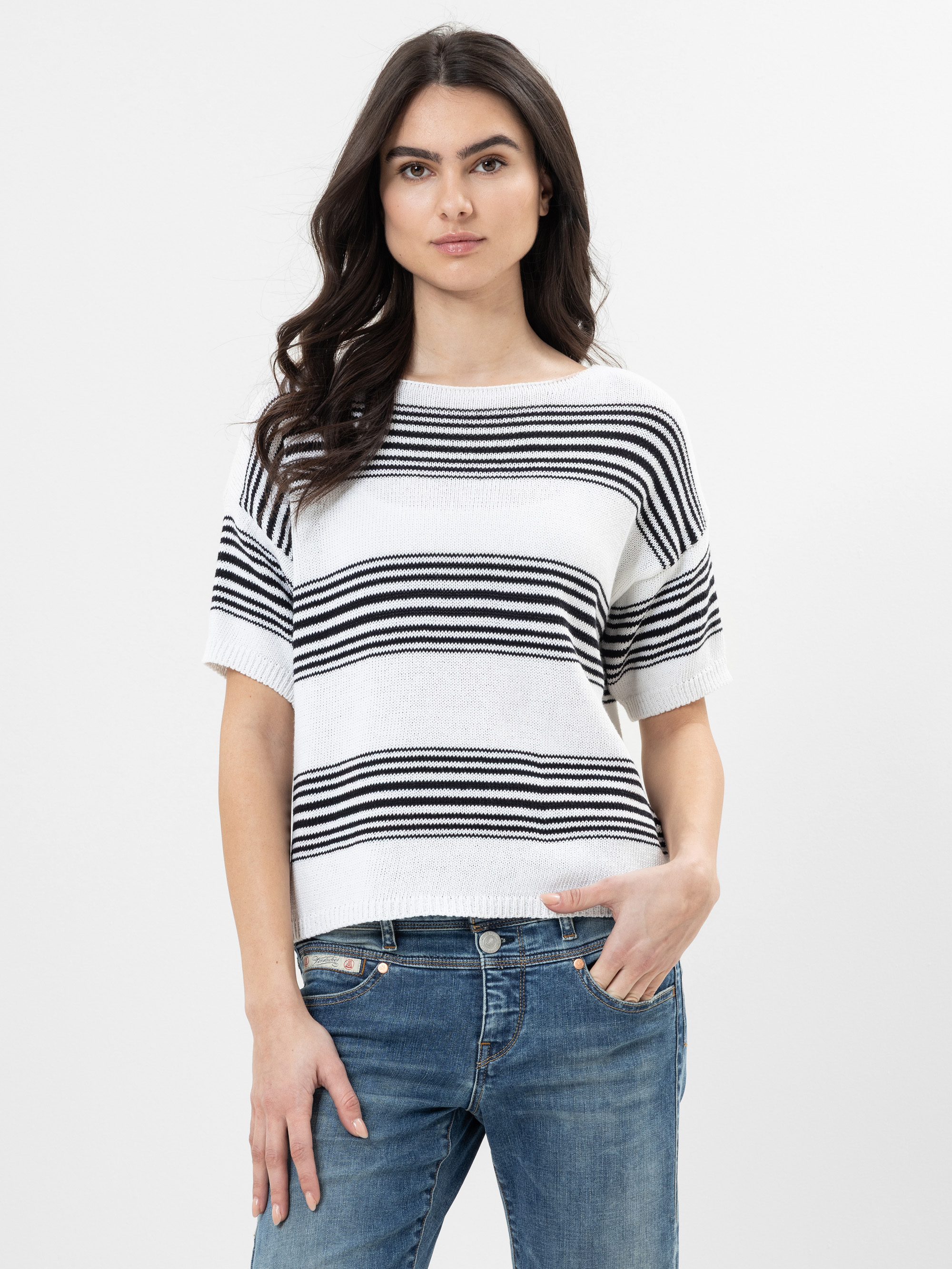 Eine Frau mit langen dunklen Haaren, die einen weißen kurzärmeligen Pullover mit schwarzen Querstreifen und blaue Jeans trägt, steht vor einem schlichten hellen Hintergrund und blickt mit neutralem Blick in die Kamera.
