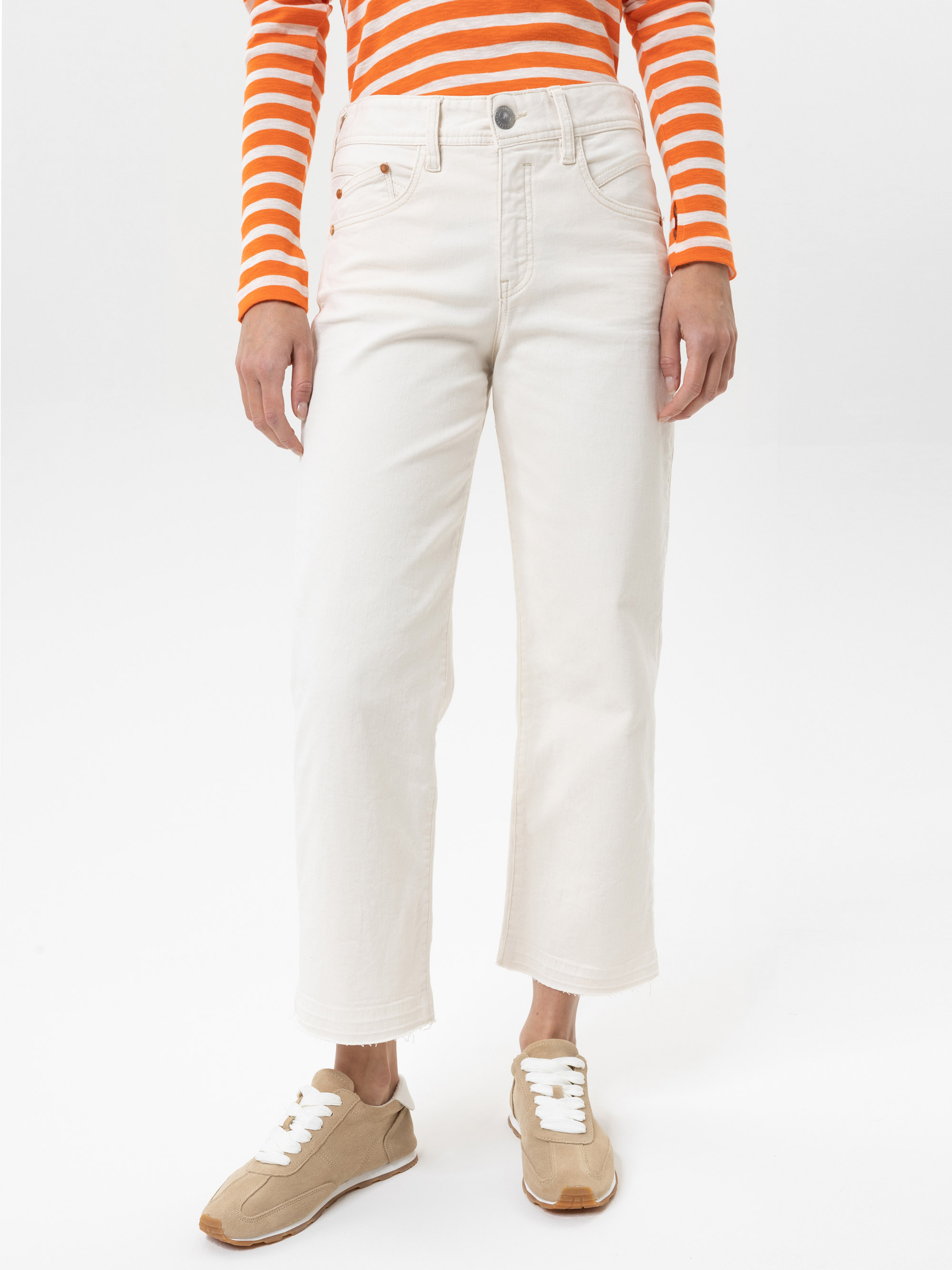 Person, die cremefarbene Jeans mit geradem Bein, beigefarbene Turnschuhe mit weißen Schnürsenkeln und ein orange-weiß gestreiftes langärmeliges Hemd trägt und vor einem schlichten weißen Hintergrund steht.