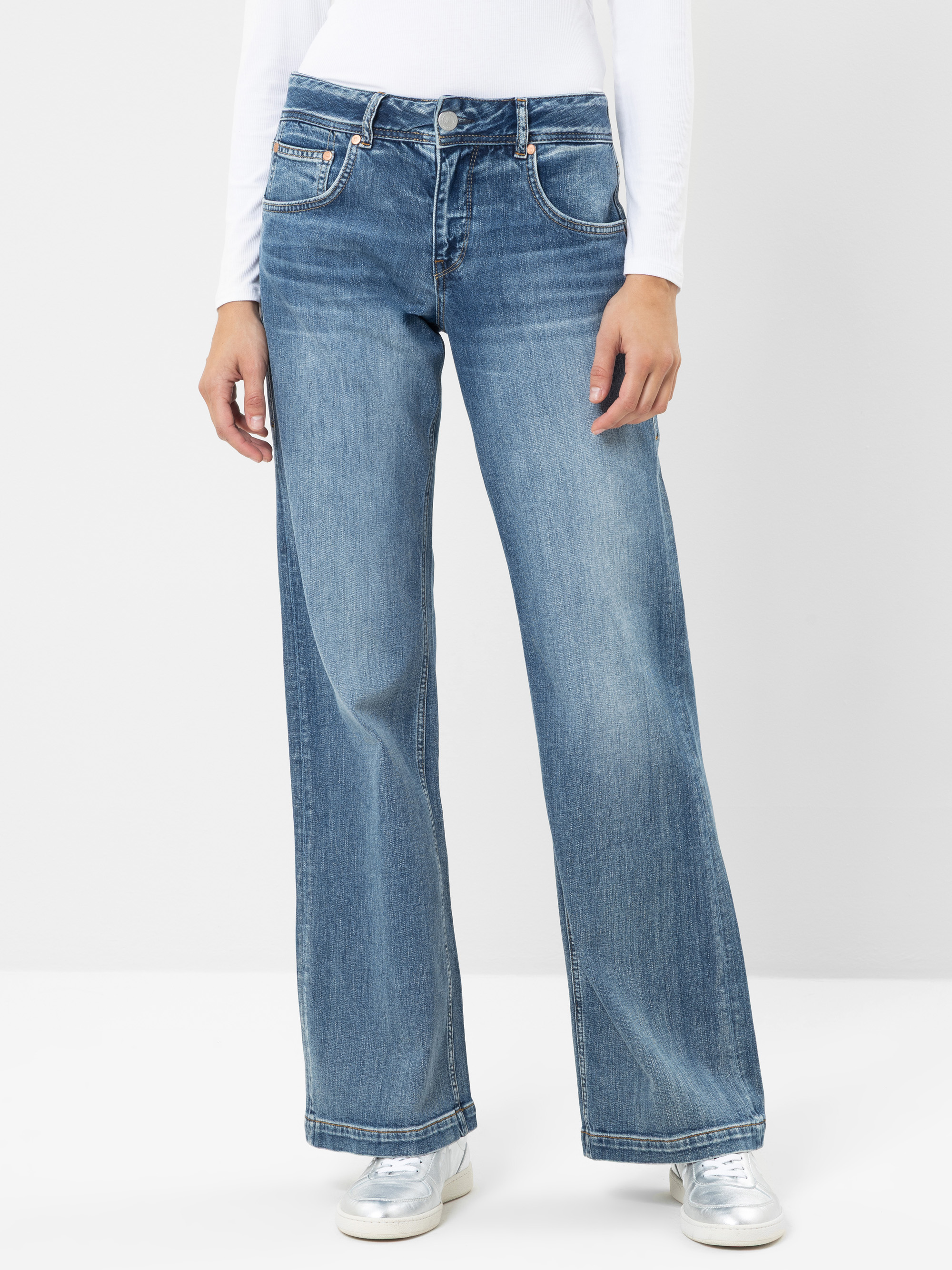 Eine Person, die ein weißes Langarmshirt, eine blaue Jeans mit weitem Bein und silberne Schuhe trägt, steht vor einem schlichten, hellen Hintergrund, die Hände entspannt an den Seiten.