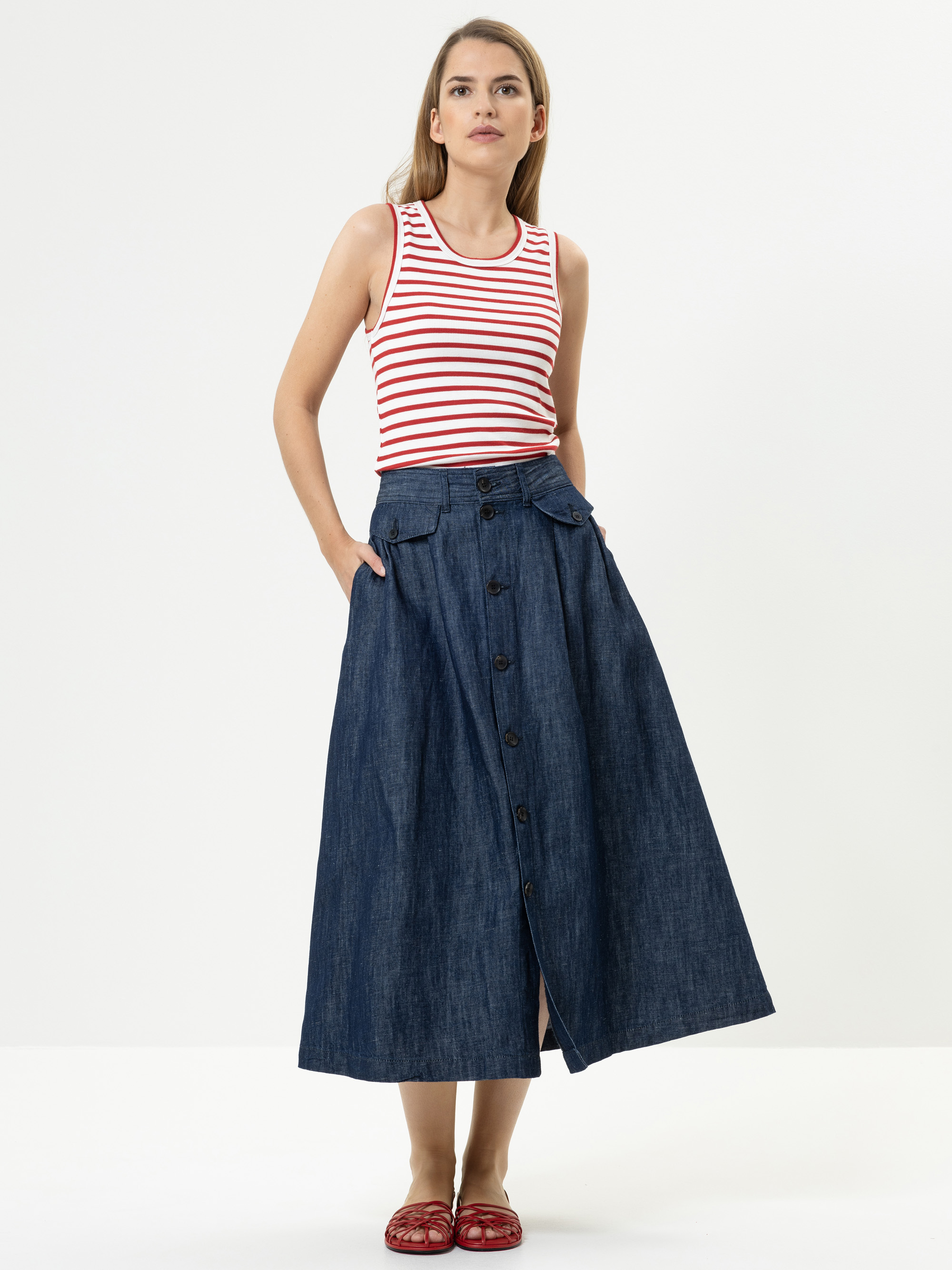 Eine Frau steht vor einem weißen Hintergrund und trägt ein ärmelloses, rot-weiß gestreiftes Oberteil, einen hochtaillierten, geknöpften Jeans-Maxirock und rote Schuhe. Ihre Hände stecken in den Taschen ihres Rocks.