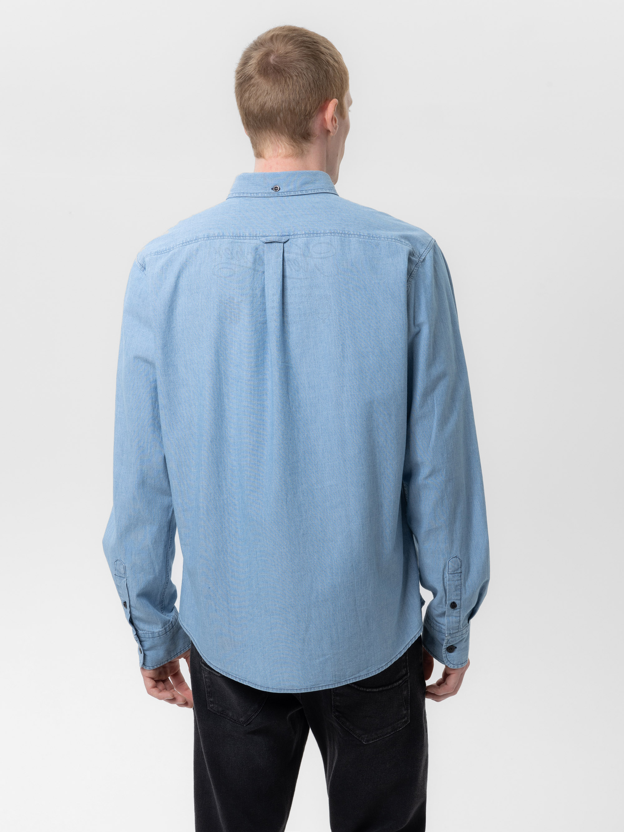 Eine Person mit kurzen hellbraunen Haaren steht mit dem Rücken zur Kamera und trägt ein hellblaues, langärmeliges Button-up-Hemd und eine schwarze Hose vor einem schlichten weißen Hintergrund.