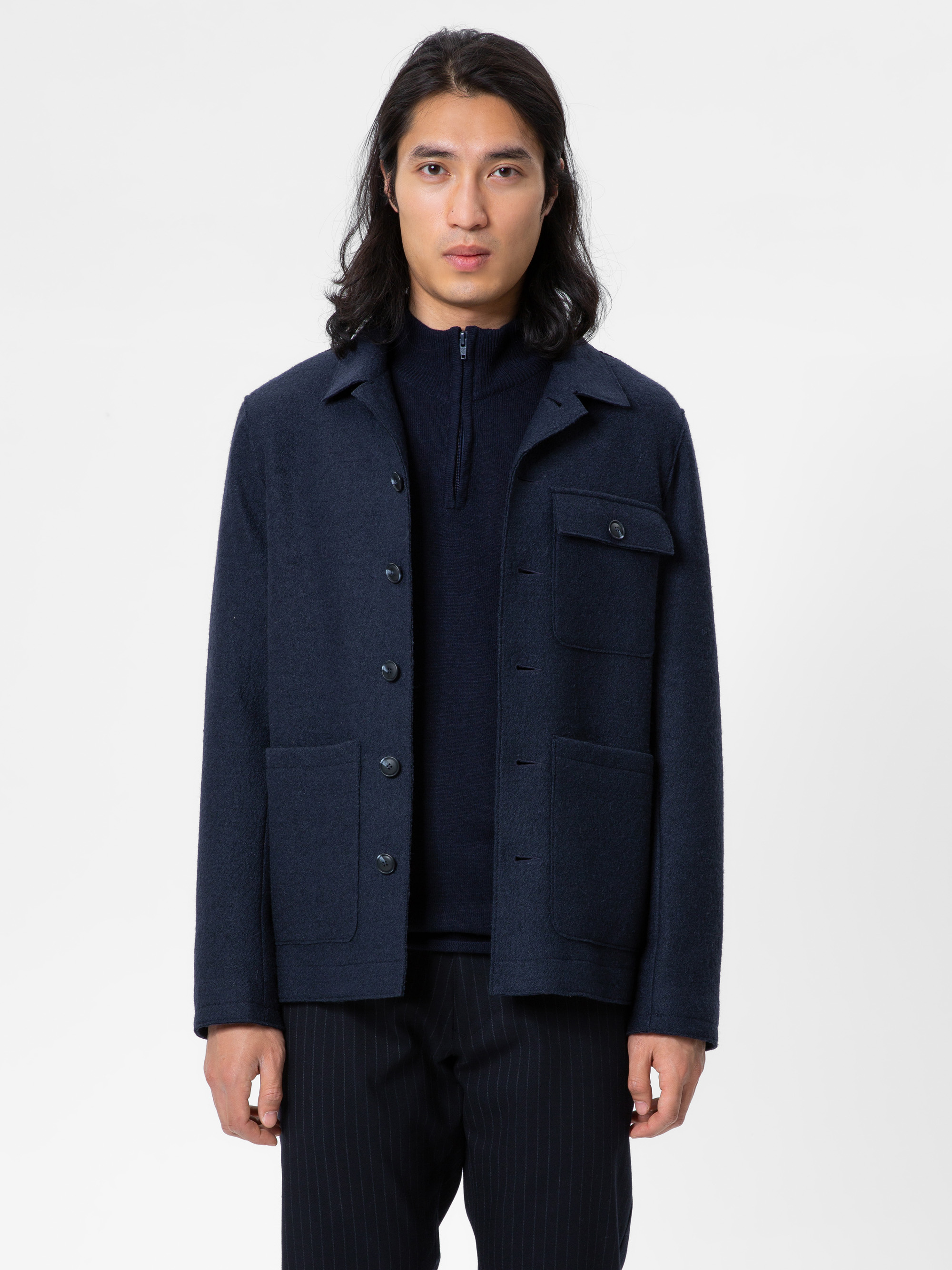 Eine Person mit langen dunklen Haaren trägt eine marineblaue Button-up-Jacke mit großen Taschen über einem dunklen Oberteil und einer dunklen Nadelstreifenhose und steht vor einem schlichten weißen Hintergrund.