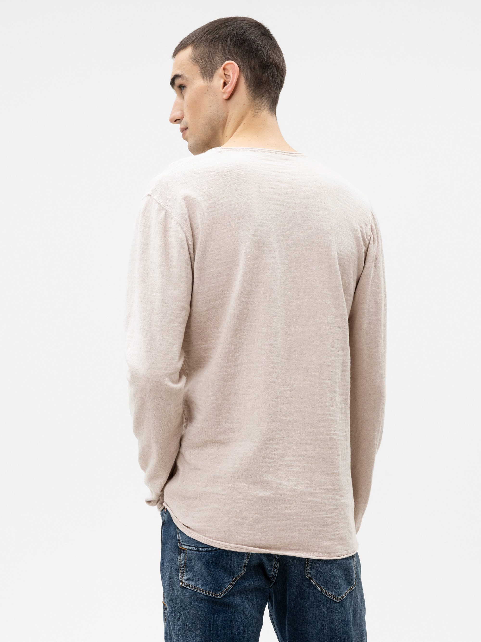 Ein Mann mit kurzen dunklen Haaren, der ein hellbeiges langärmeliges Hemd und eine blaue Jeans trägt, steht mit dem Rücken zur Kamera vor einem schlichten weißen Hintergrund.