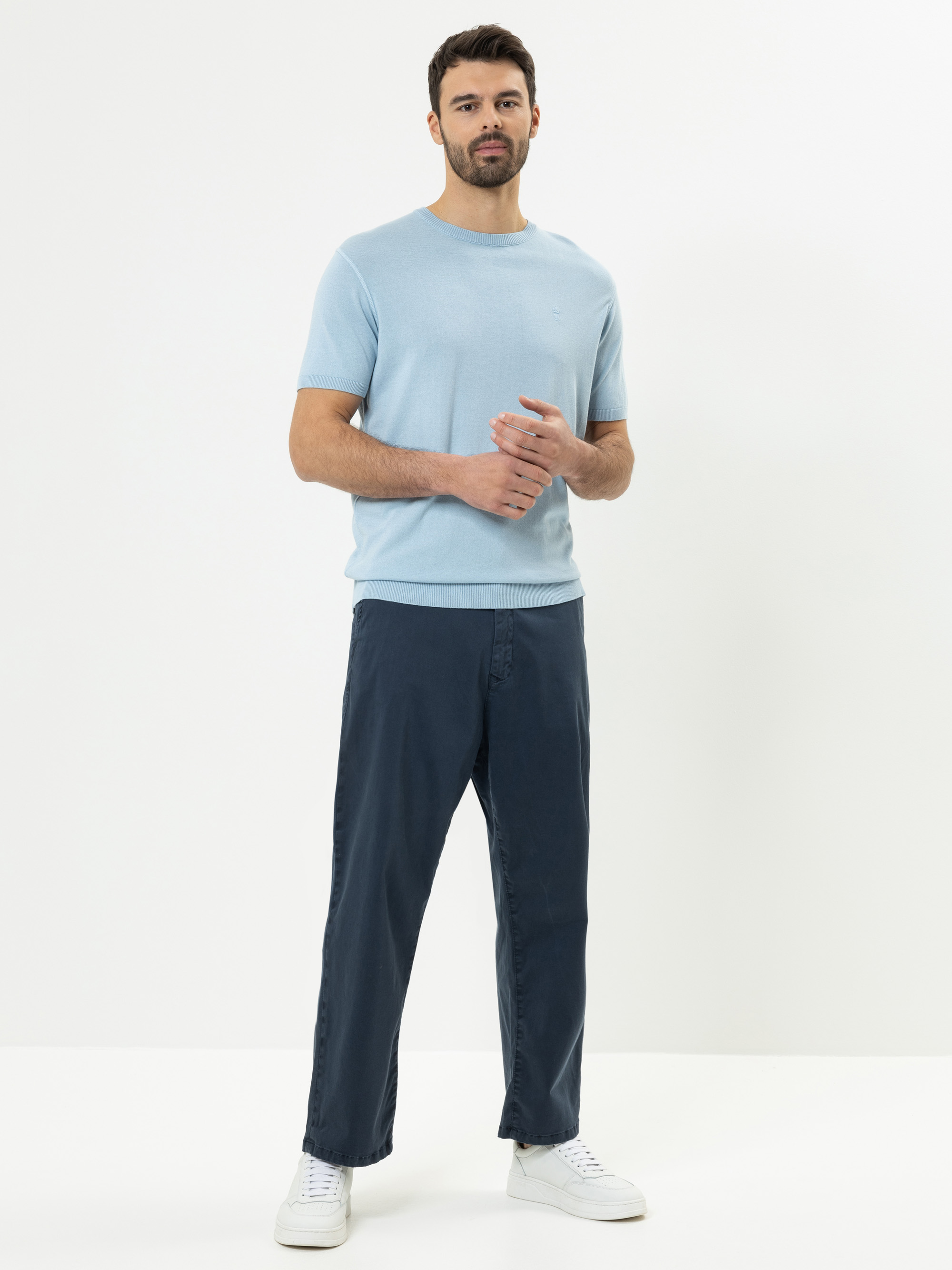 Ein Mann steht vor einem schlichten weißen Hintergrund und trägt ein hellblaues kurzärmeliges Hemd, eine dunkelblaue Hose und weiße Turnschuhe. Er hat kurzes dunkles Haar, einen Bart und seine Hände sind leicht vor ihm verschränkt.