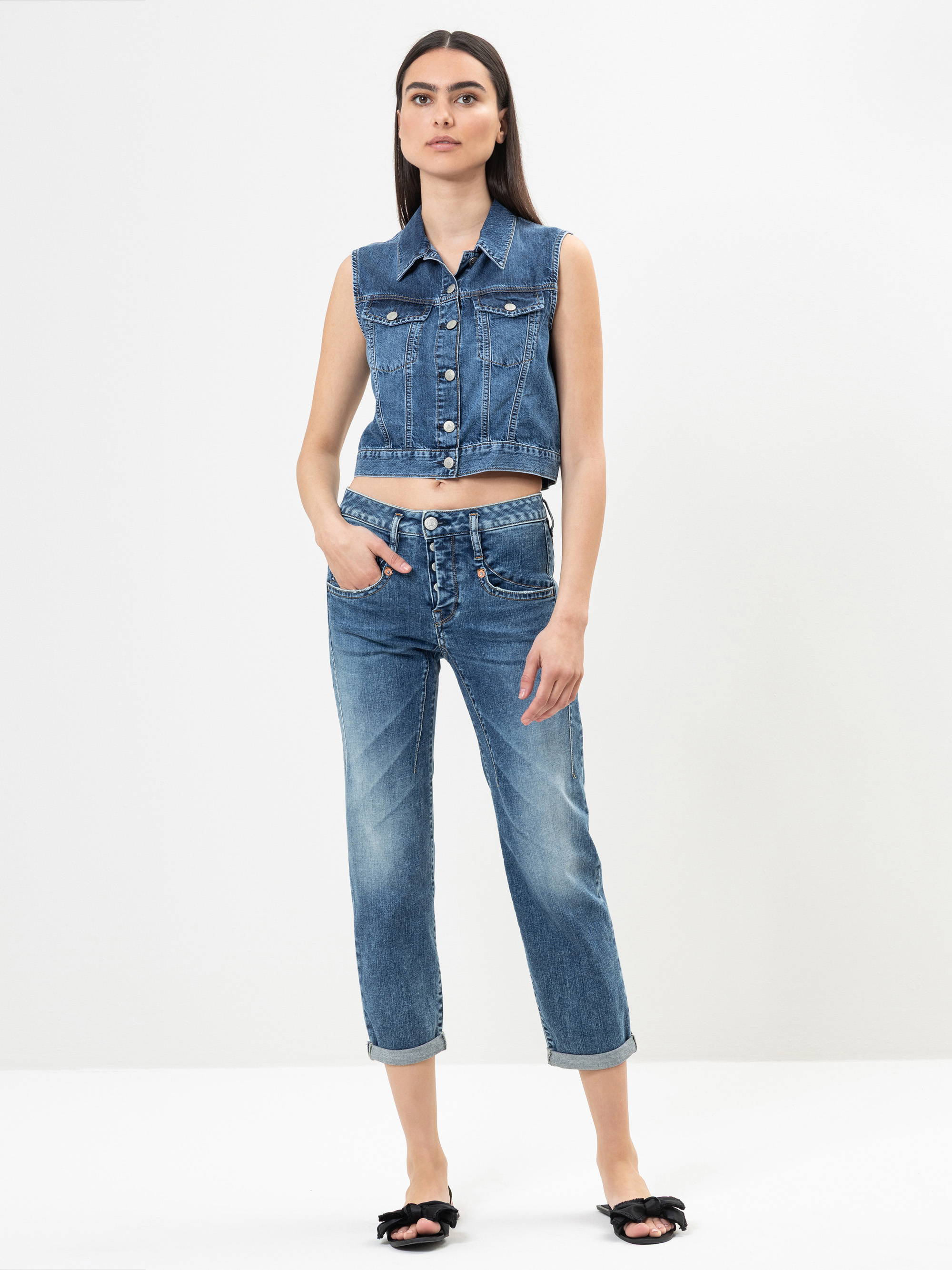 Eine Frau steht vor einem weißen Hintergrund und trägt eine ärmellose Jeansweste, blaue Jeans mit Bündchen und schwarze Schleifensandalen. Sie hat langes dunkles Haar und eine Hand in der Tasche.