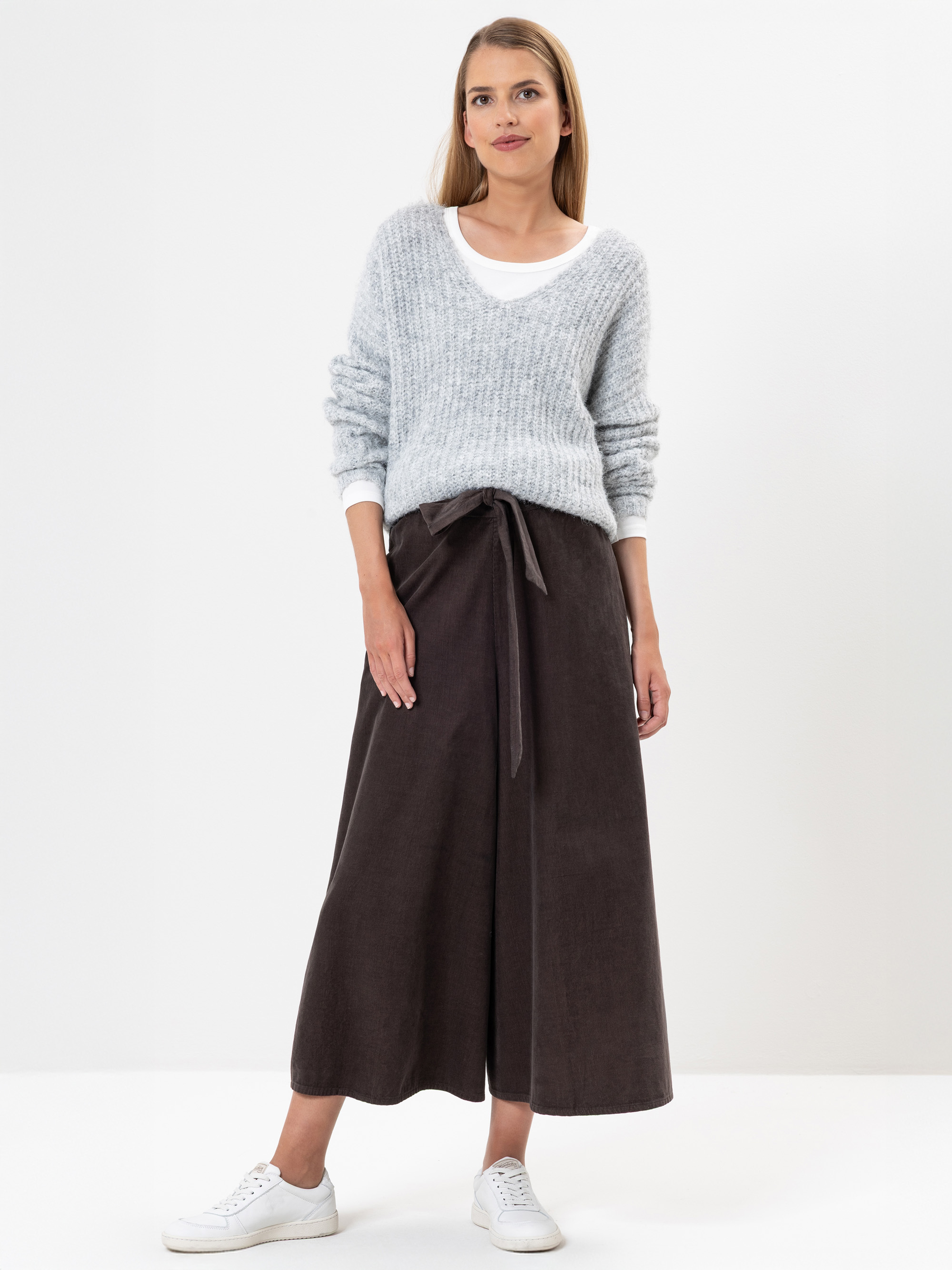 Eine Frau steht vor einem weißen Hintergrund und trägt einen hellgrauen Strickpullover über einem weißen Oberteil, einen langen braunen Rock mit Bindegürtel und weiße Turnschuhe. Sie hat glattes, hellbraunes Haar und einen neutralen Gesichtsausdruck
