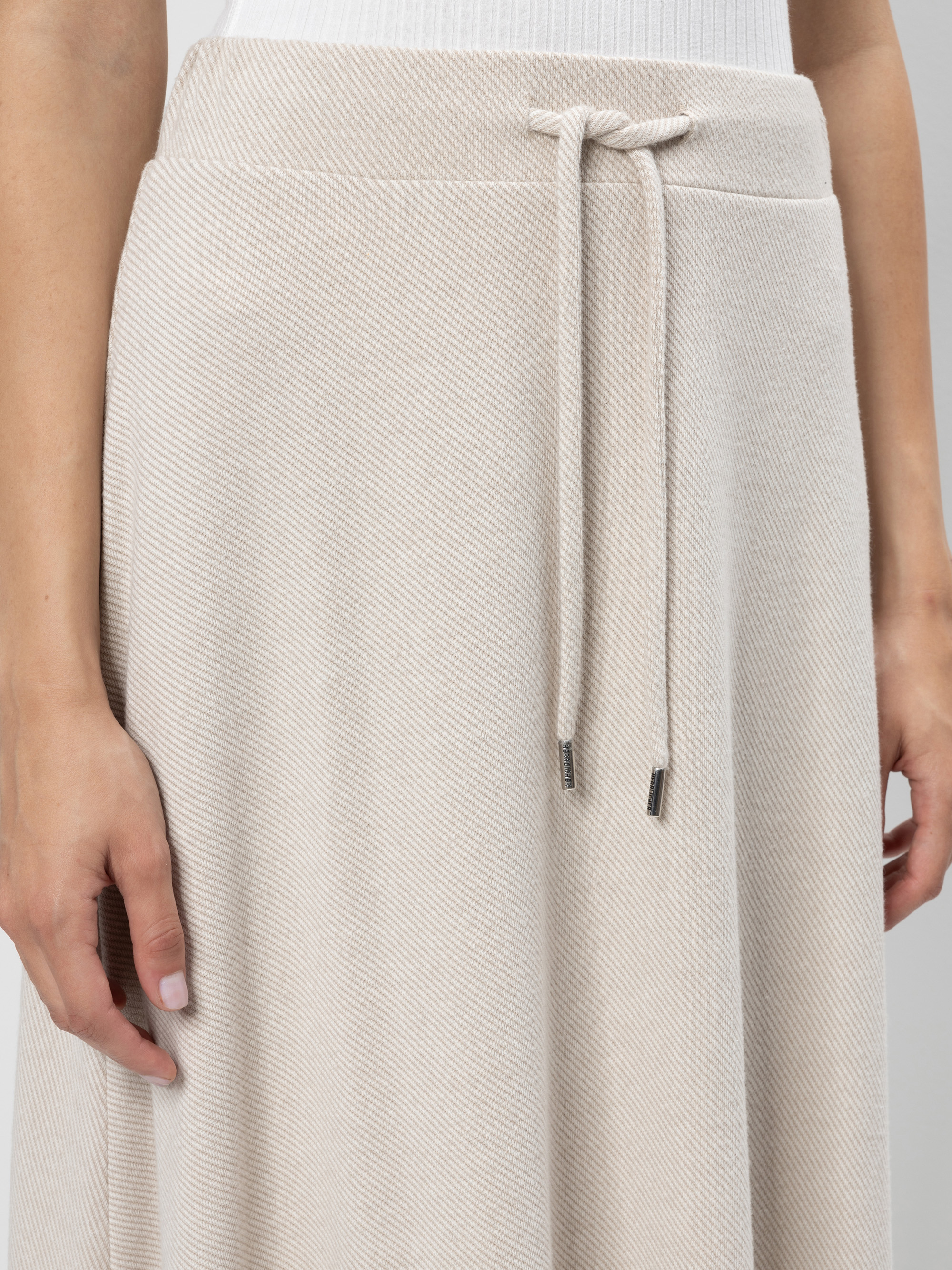Nahaufnahme einer Person, die einen hellbeigen, strukturierten Rock mit Kordelzug in der Taille und Metallspitzen trägt, kombiniert mit einem weißen Oberteil. Man sieht die linke Hand der Person, die an ihrer Seite ruht.