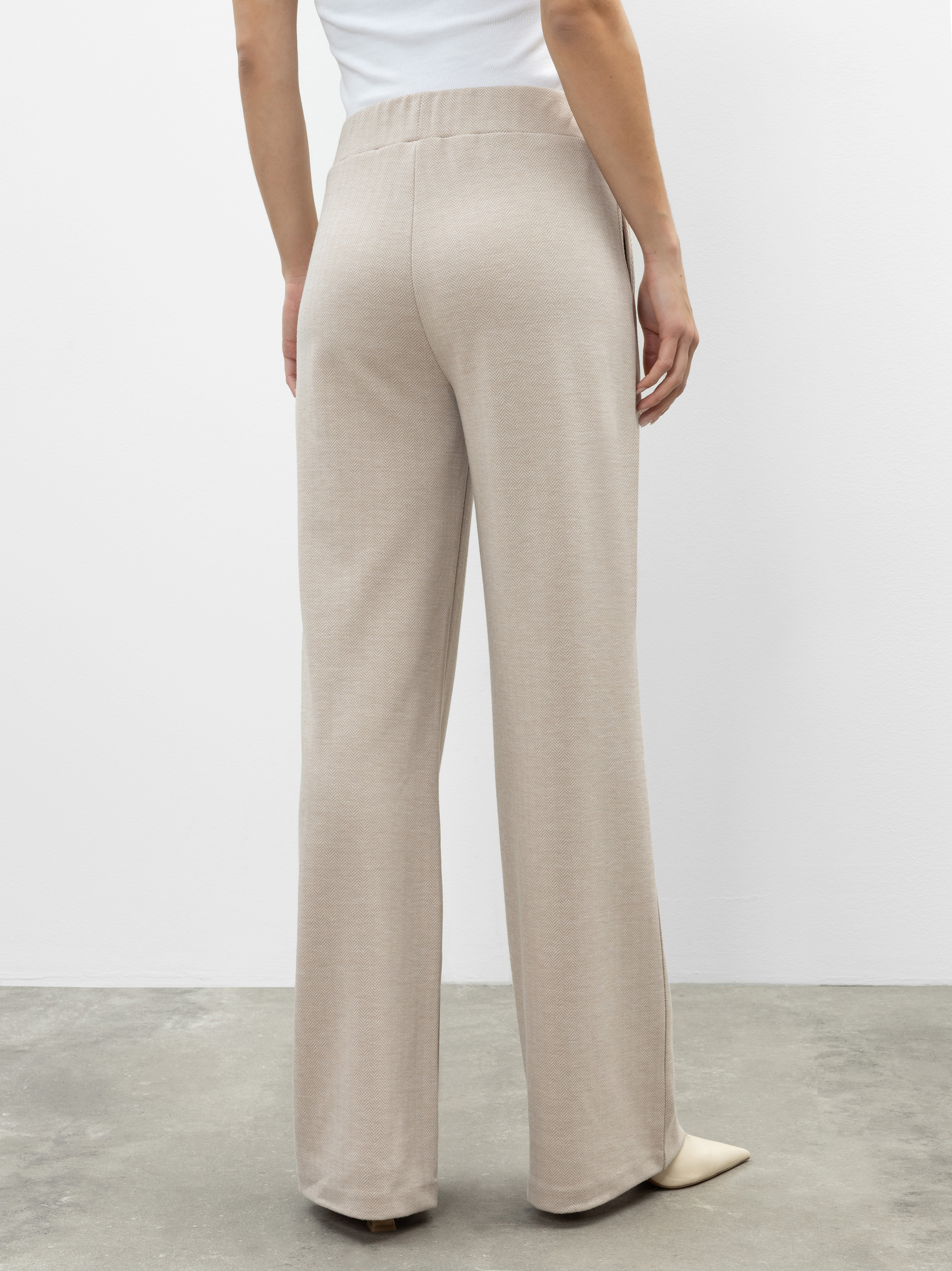 Eine Person, die eine hochtaillierte, locker sitzende beigefarbene Hose und ein weißes Oberteil trägt, steht auf einem grauen Betonboden, den Blick von der Kamera abgewandt, und hält die linke Hand an die Seite gelegt.