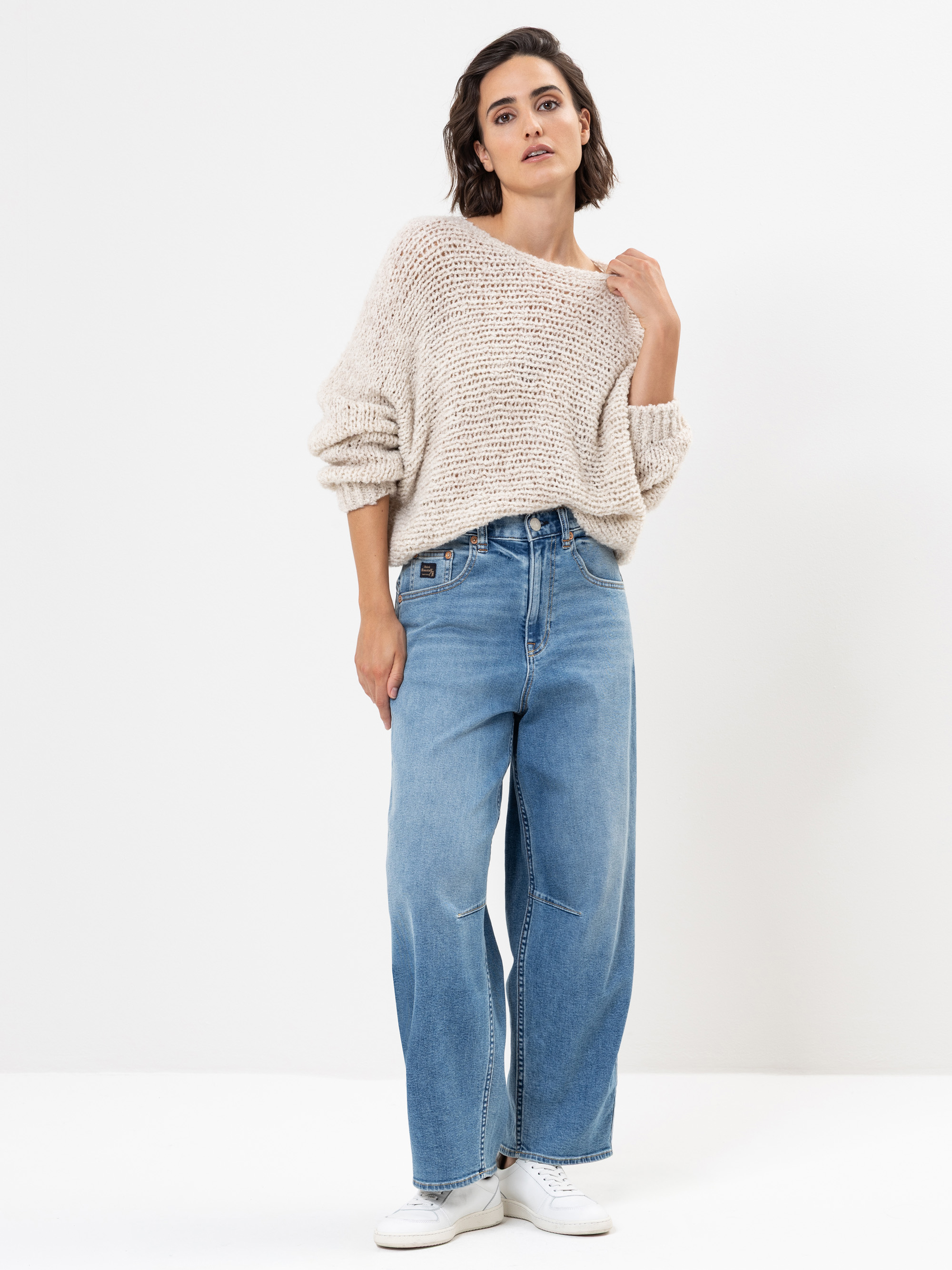 Eine Frau steht vor einem weißen Hintergrund und trägt einen lockeren beigefarbenen Strickpullover, lässige Bluejeans und weiße Turnschuhe. Sie hat kurzes dunkles Haar und einen neutralen Gesichtsausdruck, wobei eine Hand ihren Pullover berührt.