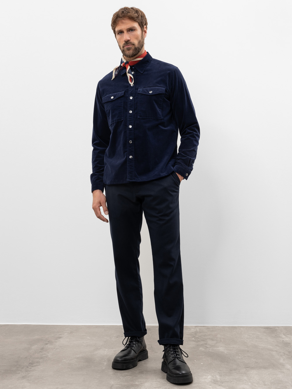 Ein Mann steht vor einem schlichten weißen Hintergrund, trägt ein marineblaues Button-up-Hemd, eine schwarze Hose, schwarze Schnürschuhe und einen rot gemusterten Schal, der um seinen Hals gebunden ist.