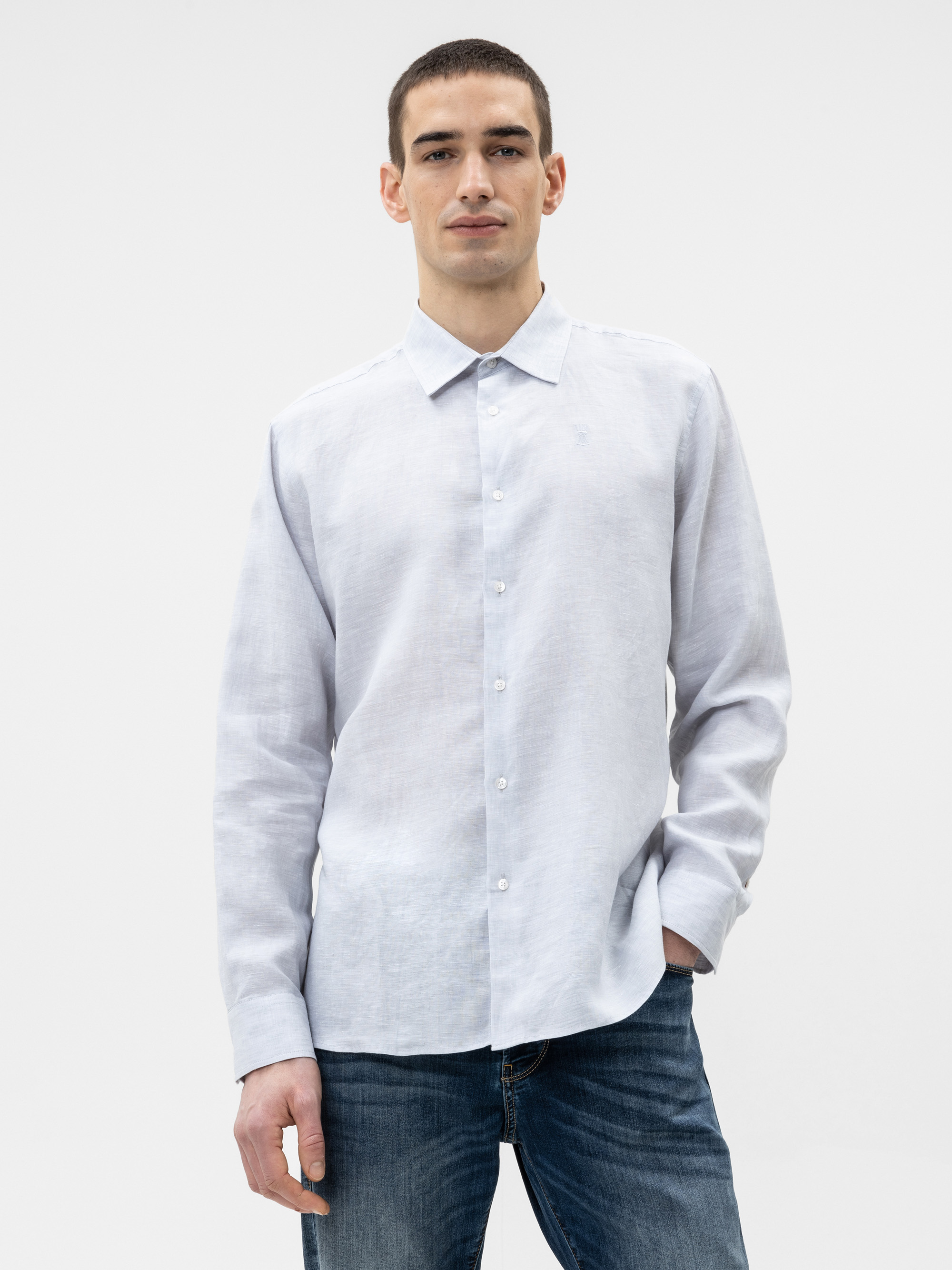 Ein Mann in einem hellgrauen, langärmeligen Button-up-Hemd und blauen Jeans steht vor einem schlichten weißen Hintergrund und hat eine Hand in der Tasche.