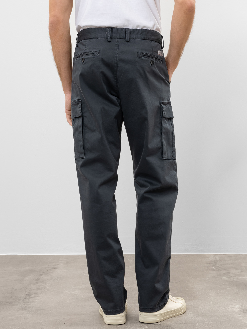 Eine Person, die eine dunkelgraue Cargohose und weiße Turnschuhe trägt, steht mit dem Gesicht zur Kamera und zeigt die Rückansicht der Hose, die Klappentaschen und seitliche Cargotaschen hat.