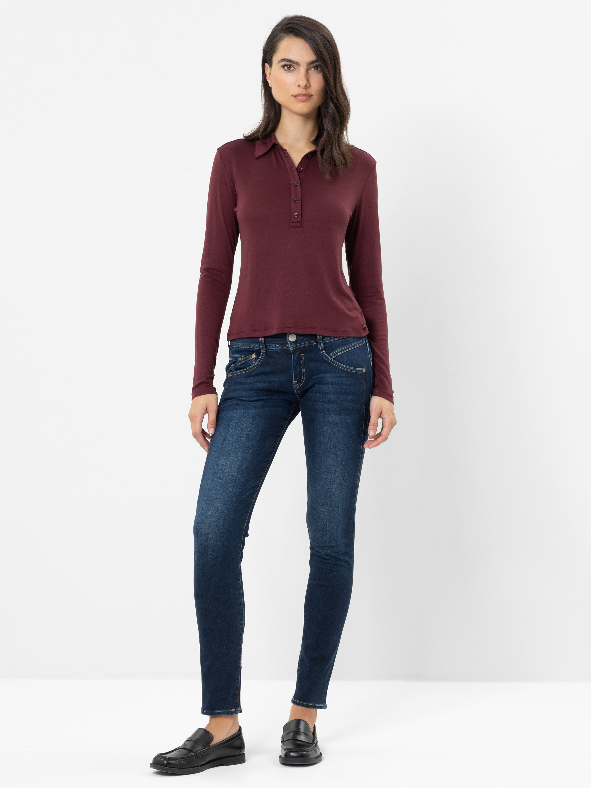 Eine Frau steht vor einem weißen Hintergrund und trägt ein burgunderrotes Poloshirt mit langen Ärmeln, dunkelblaue Skinny-Jeans und schwarze Slipper.