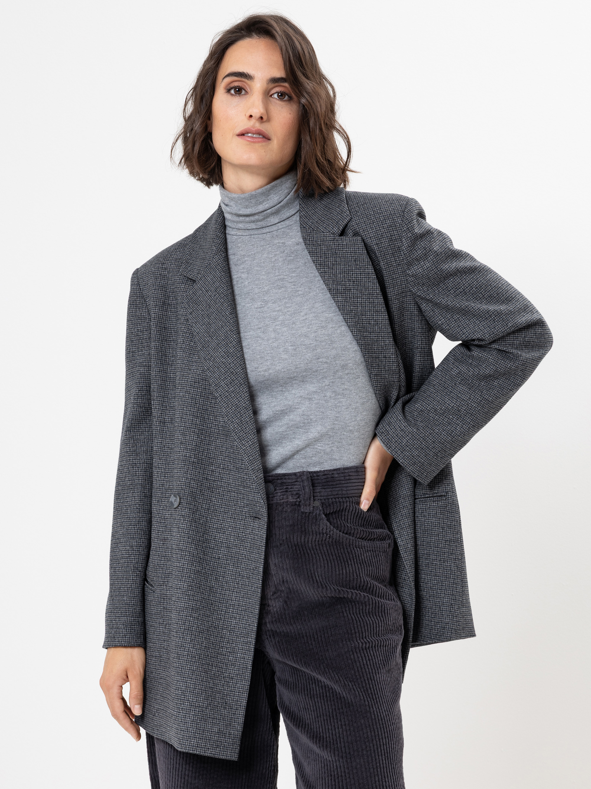 Eine Frau mit kurzen braunen Haaren trägt einen grauen Rollkragenpullover, einen übergroßen grauen Blazer und eine dunkelgraue Cordhose. Sie steht vor einem schlichten weißen Hintergrund und hat einen neutralen Gesichtsausdruck.