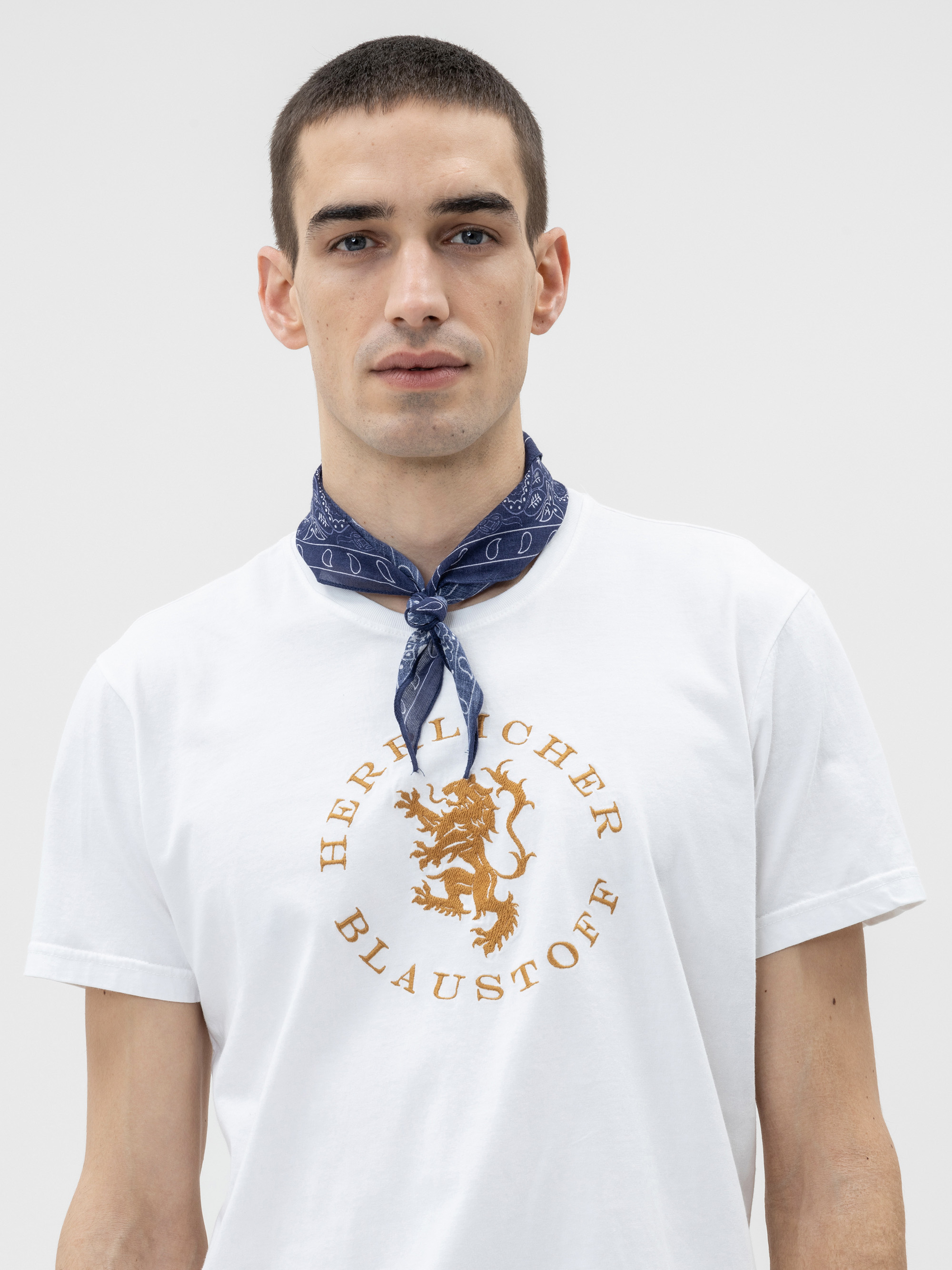Ein junger Mann mit kurzen dunklen Haaren trägt ein weißes T-Shirt mit einem goldenen Löwenemblem und der Aufschrift Herrlicher Blaustoff. Er hat ein blau gemustertes Kopftuch um den Hals gebunden und steht vor einem schlichten hellen Hintergrund.
