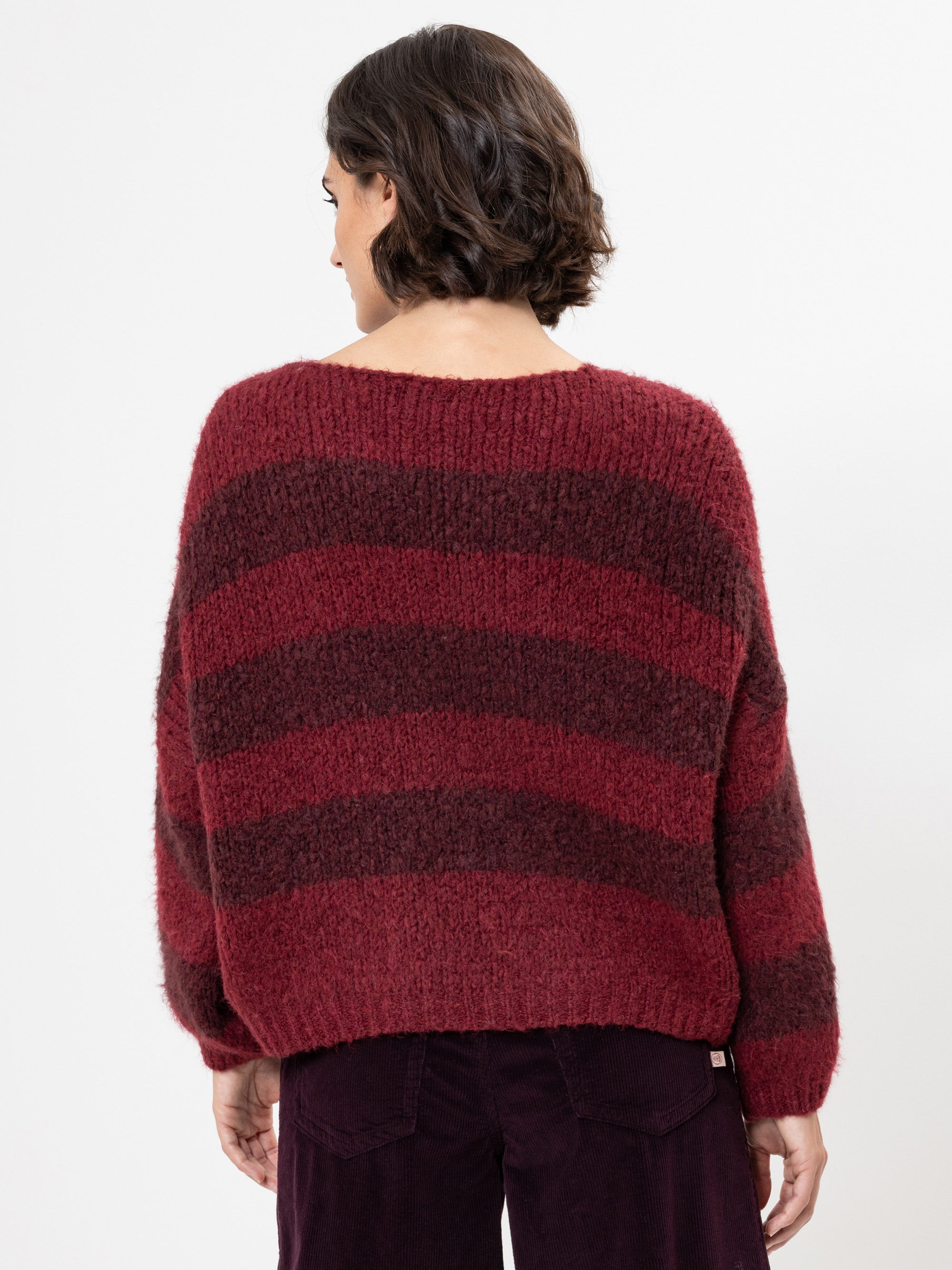 Eine Person mit kurzen, gewellten braunen Haaren wird von hinten gezeigt. Sie trägt einen lockeren, gestrickten Pullover mit breiten kastanienbraunen und dunkelroten Streifen und eine dunkle Hose. Der Hintergrund ist einfarbig weiß.